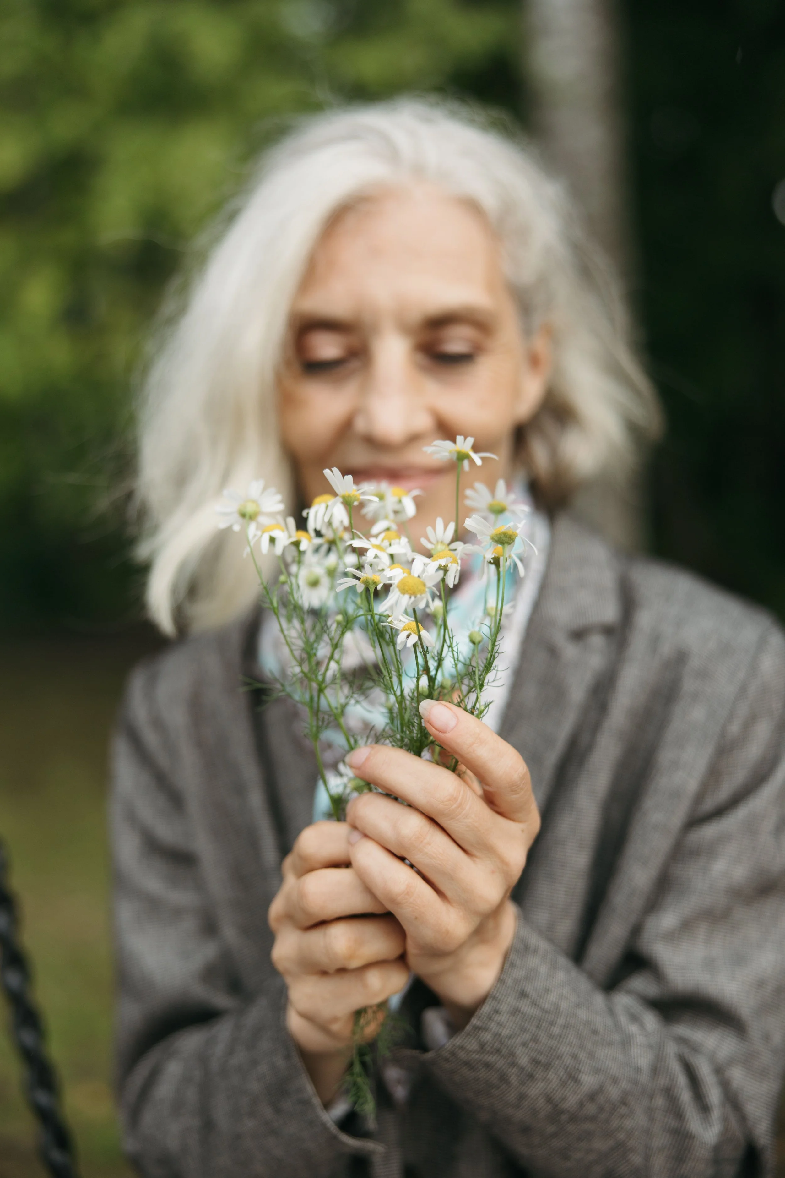 A woman with silver hair smiling and holding a bouquet of small white daisies outdoors.