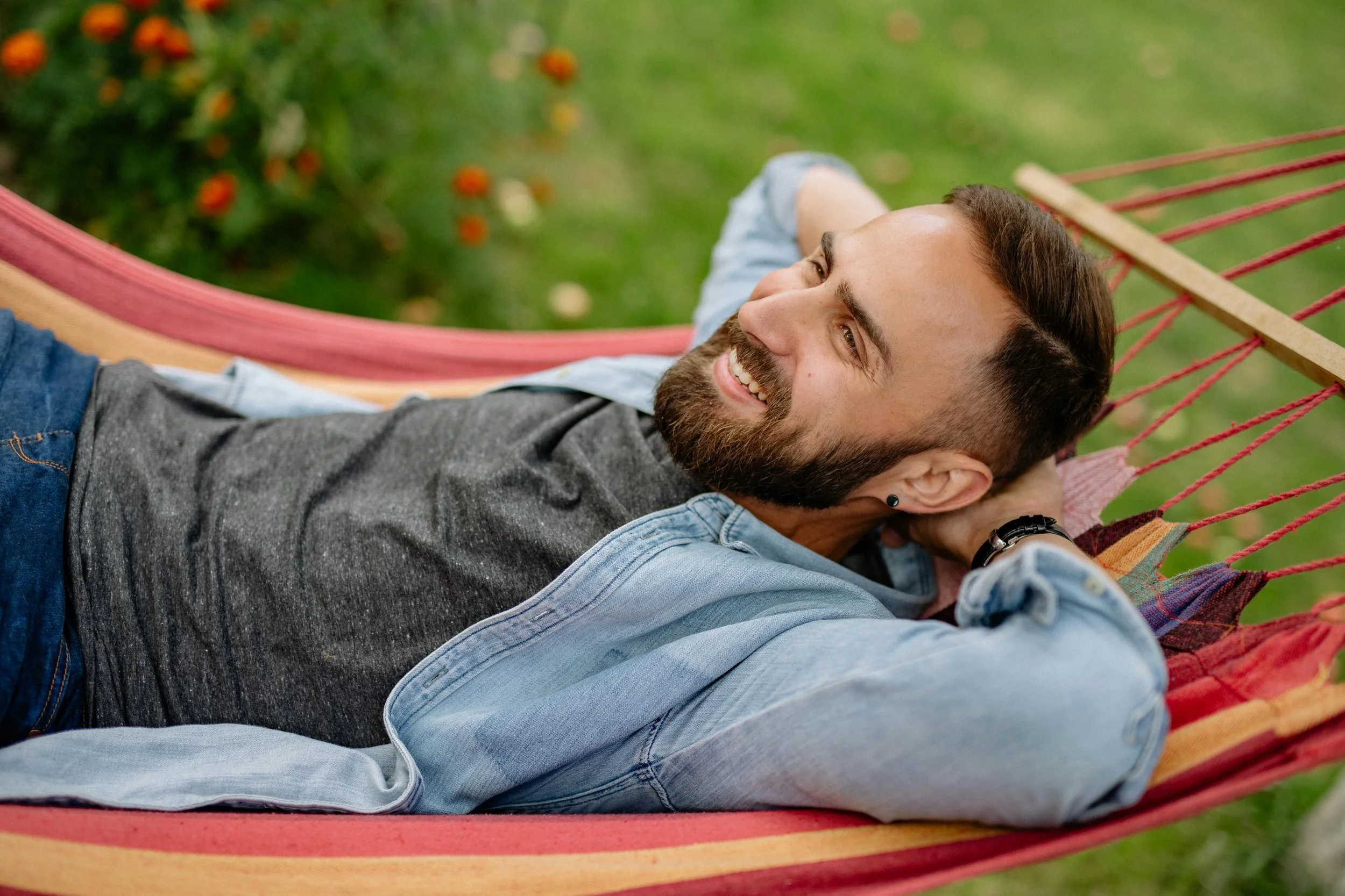 Man with a beard relaxing on a hammock outdoors, smiling, with grass and flowers in the background.