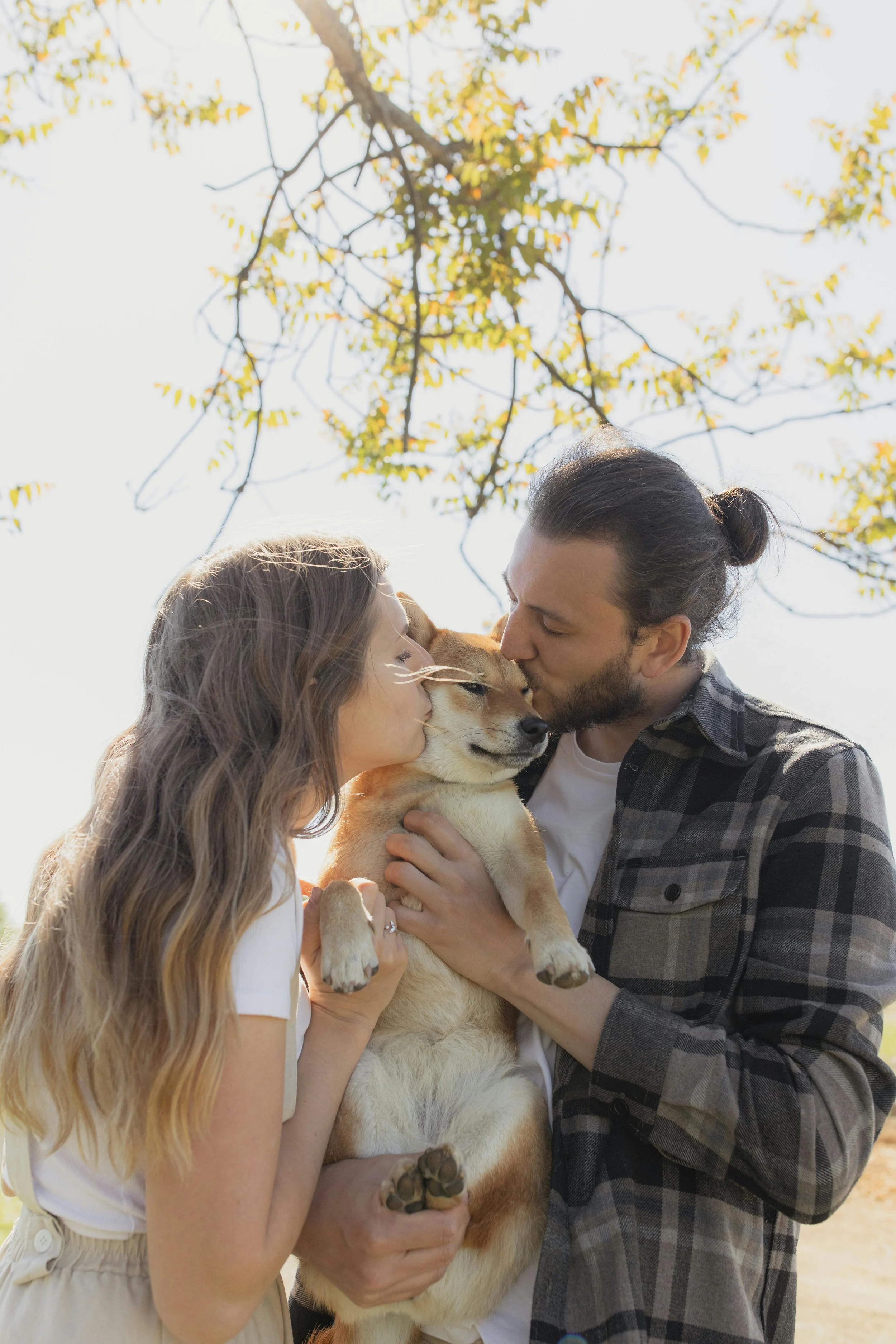 A young girl, a man, and a dog sharing a kiss outdoors under a tree with yellow leaves.