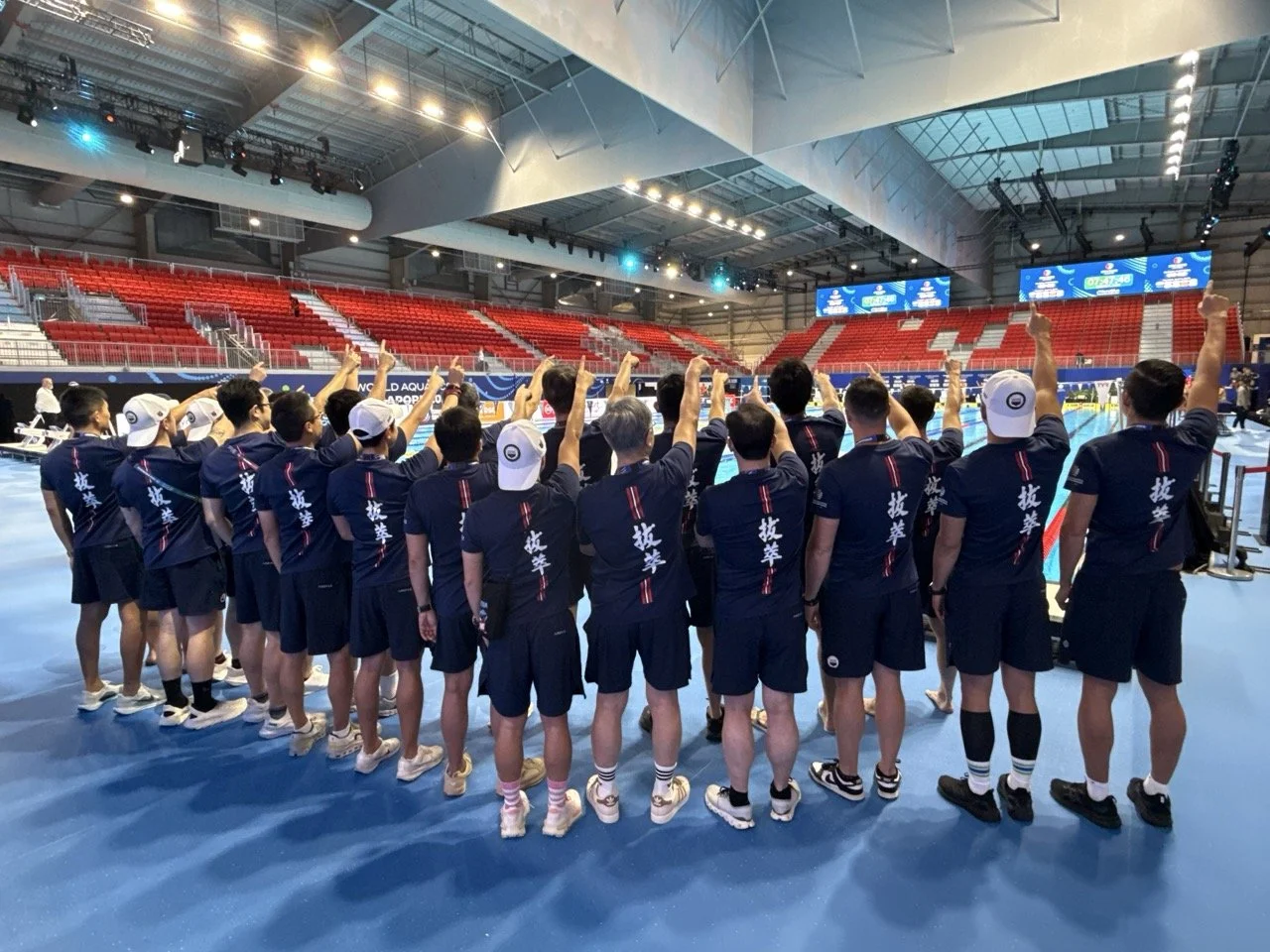 A team of young athletes standing in a sports arena, wearing navy blue uniforms with white hats, raising their right hands, inside a large indoor space with red bleachers and digital screens.