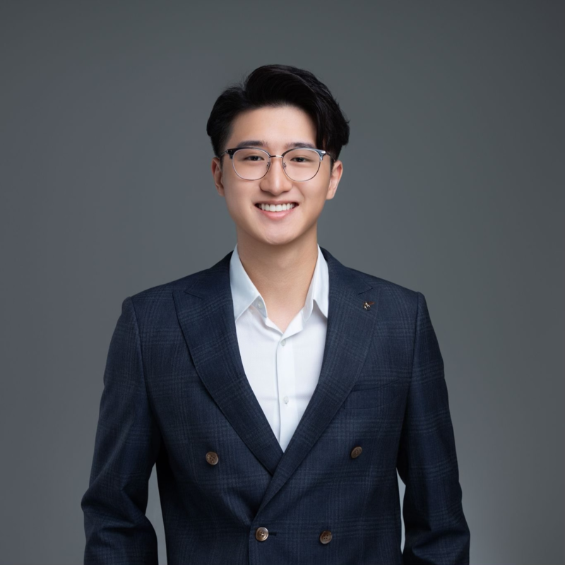 A young man in a dark blue plaid blazer and white shirt, smiling against a gray background.