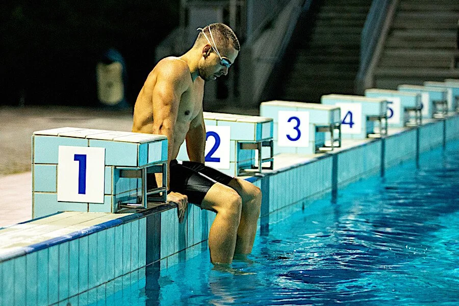 Male swimmer sitting on the edge of a swimming pool, preparing for a race, with starting blocks numbered 1 through 5 in the background.