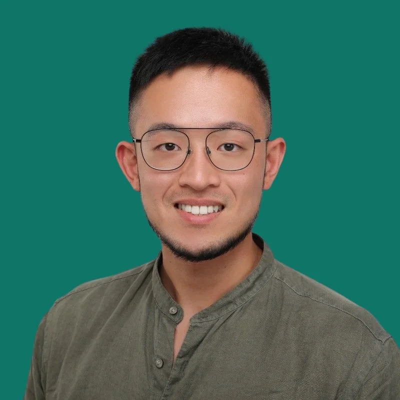 Portrait of a young Asian man with short black hair, glasses, smiling, wearing a green button-up shirt, against a solid green background.
