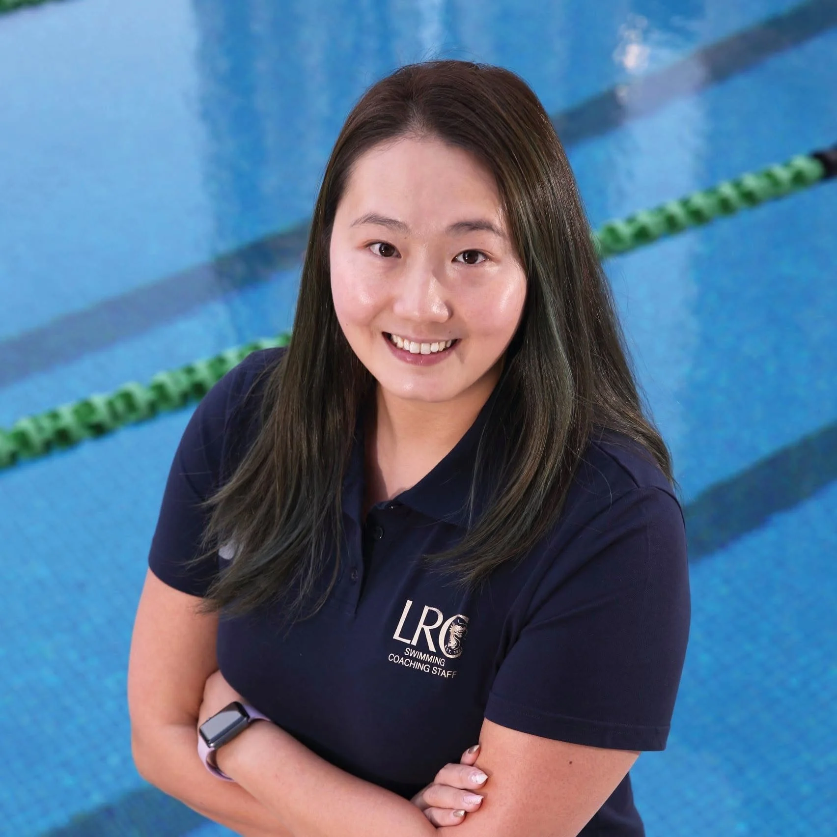 A smiling woman with long brown hair wearing a navy blue polo shirt with a logo, standing at an indoor swimming pool with blue water and lane markers.