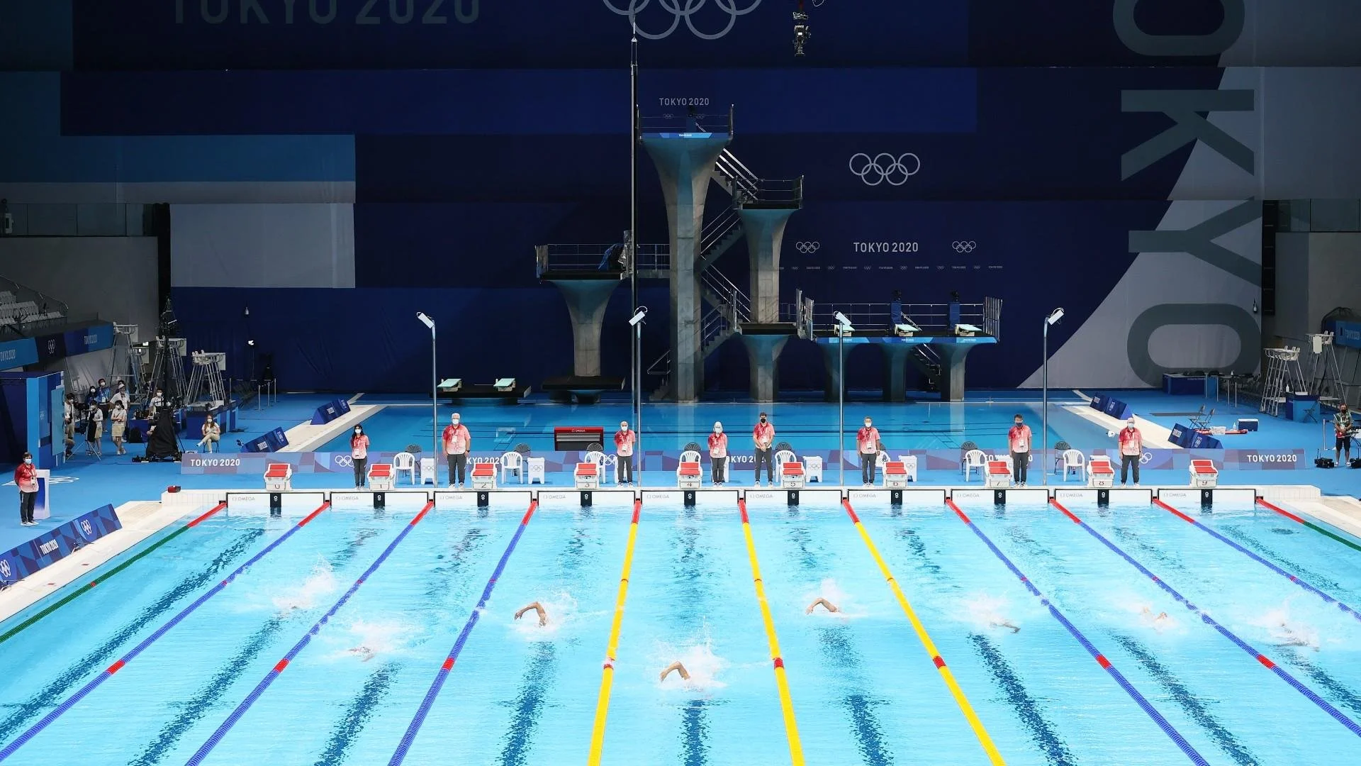 An indoor Olympic swimming pool during the Tokyo 2020 Olympics, with swimmers in the water, officials, and photographers on the pool deck, and diving boards in the background.
