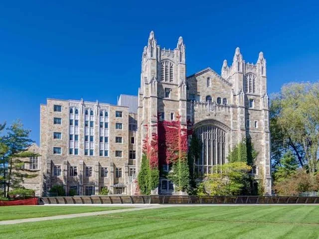 Gothic-style church or cathedral with stone walls, tall windows, and twin towers, adjacent to a modern multi-story building, with a green lawn and trees in the foreground under a clear blue sky.