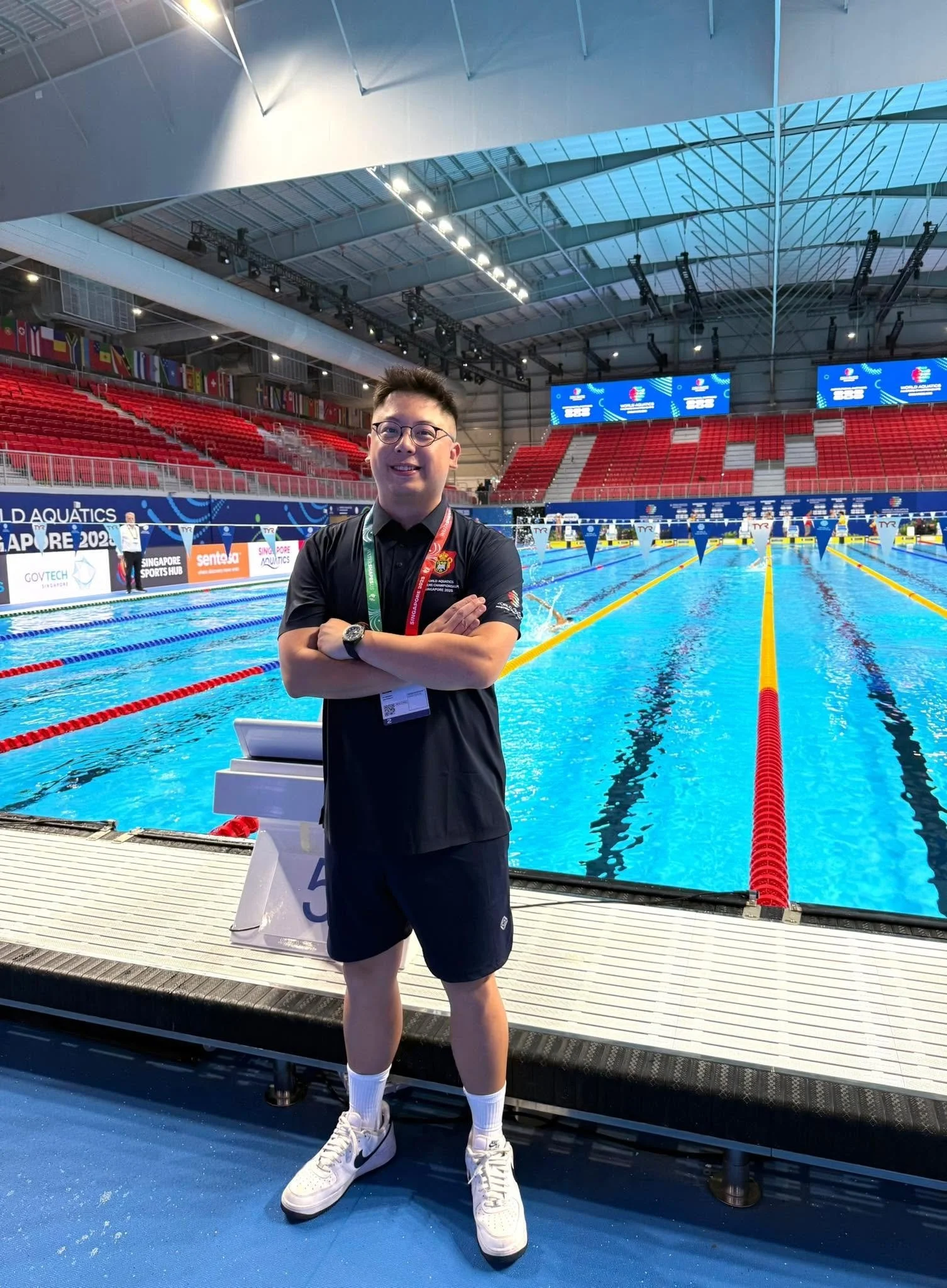 Young man in black sportswear standing with arms crossed near swimming pool in indoor aquatic center.
