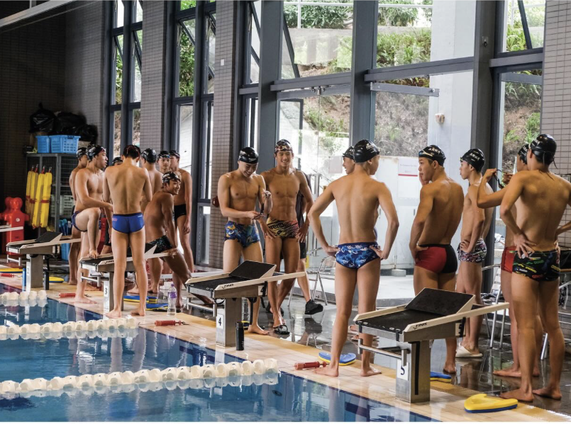 Swimmers in swim caps and swimwear preparing at starting blocks by the indoor swimming pool.