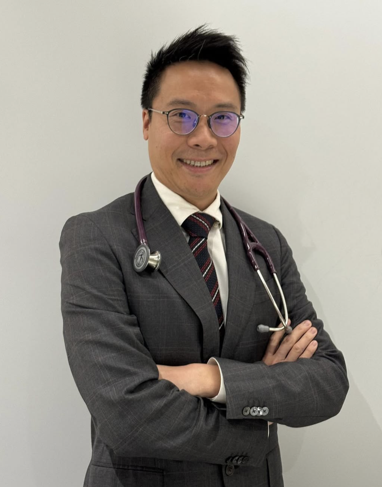 A male doctor with short black hair, wearing glasses with a slight purple tint, a dark gray suit, a white shirt, and a red and black striped tie. He has a stethoscope draped around his neck and is smiling with arms crossed. The background is plain white.