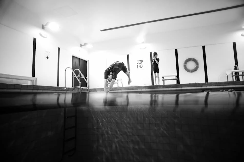 Person leaning over the edge of an indoor swimming pool, with another person taking a photo, in black and white.