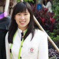 A woman with long black hair smiling outdoors, wearing a white jacket with a pink embroidered logo, in a lush garden setting.