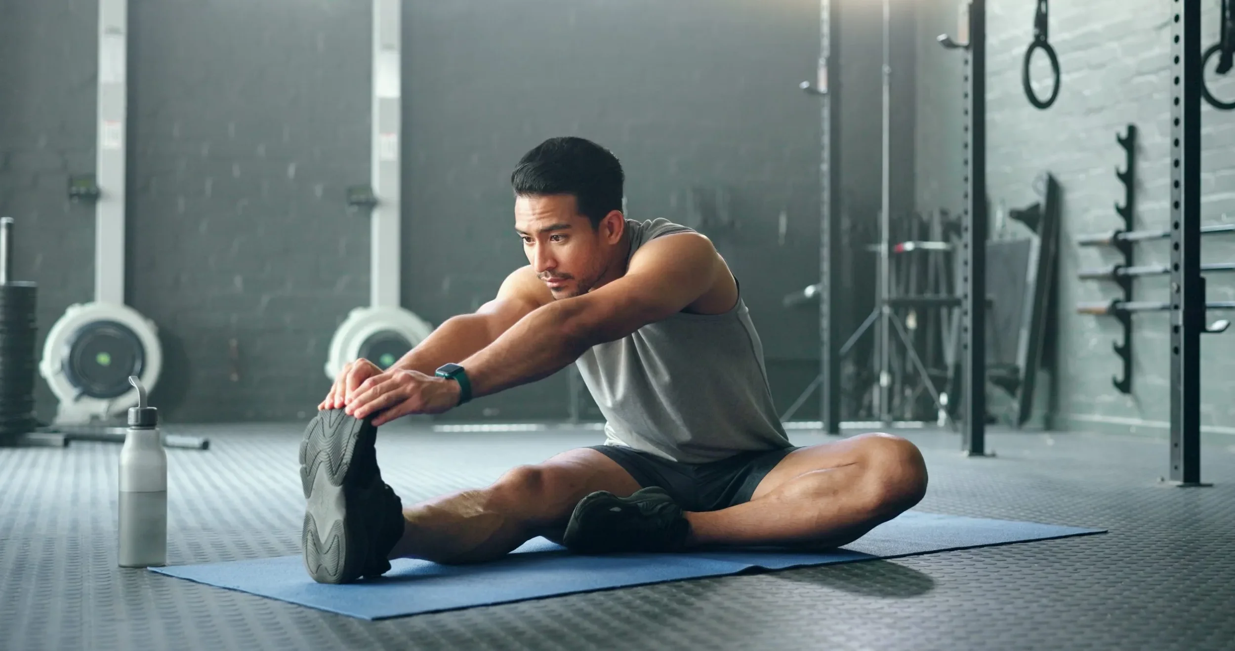 Man sitting on exercise mat in gym stretching his legs, drinking water from a bottle, with gym equipment in background.