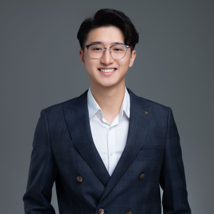 A young man with black hair and glasses smiling, wearing a navy plaid blazer and a white shirt, standing against a plain gray background.
