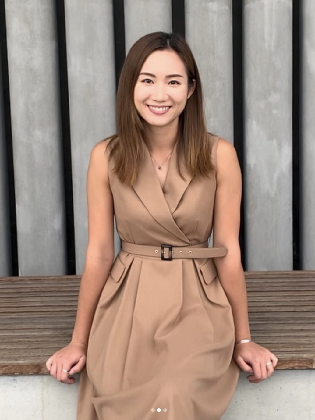 A young woman with shoulder-length brown hair, smiling, wearing a tan sleeveless dress with a belt, sitting on a wooden bench in front of a concrete and metal fence.