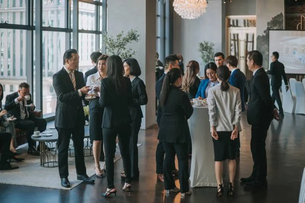 Business professionals at a networking event in a modern, well-lit conference room with large windows and chessboard decor.