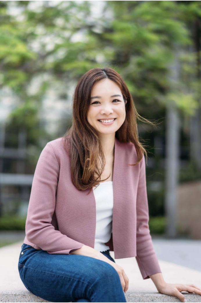 A young woman with long brown hair smiling outdoors, wearing a pink blazer and white top, sitting on a concrete ledge with greenery in the background.