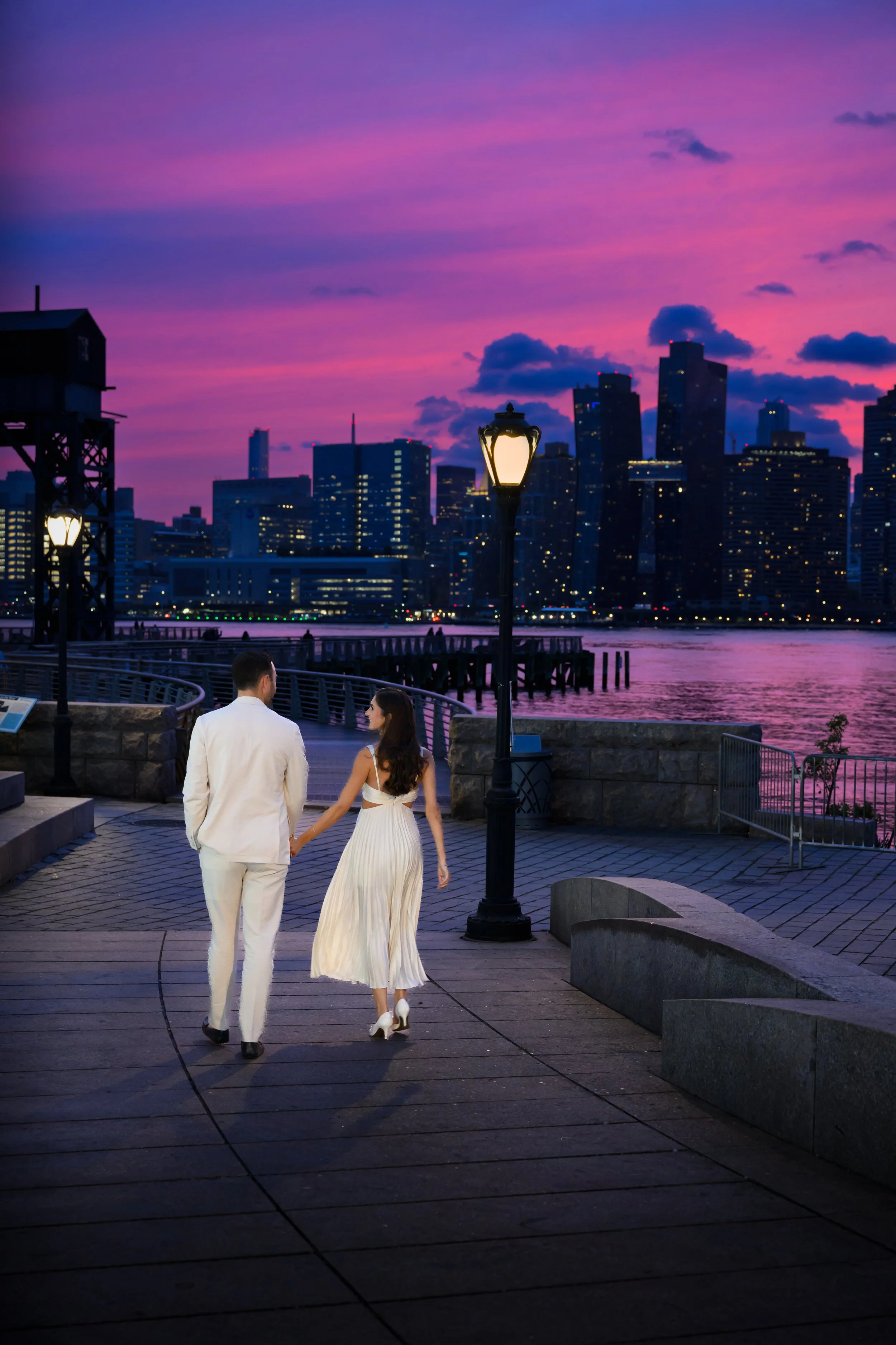 A couple in wedding attire holding hands and walking along a waterfront promenade at dusk, with city skyline and purple-pink sunset sky in the background.