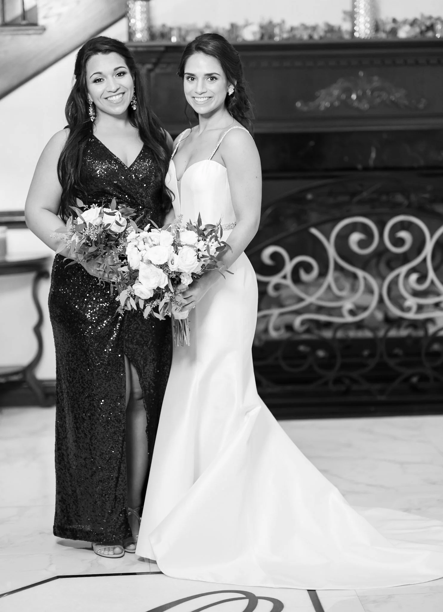 Two women in formal attire, one in a black sequined dress and the other in a white gown, smiling and holding a bouquet of flowers, standing indoors near a decorative wrought iron gate.