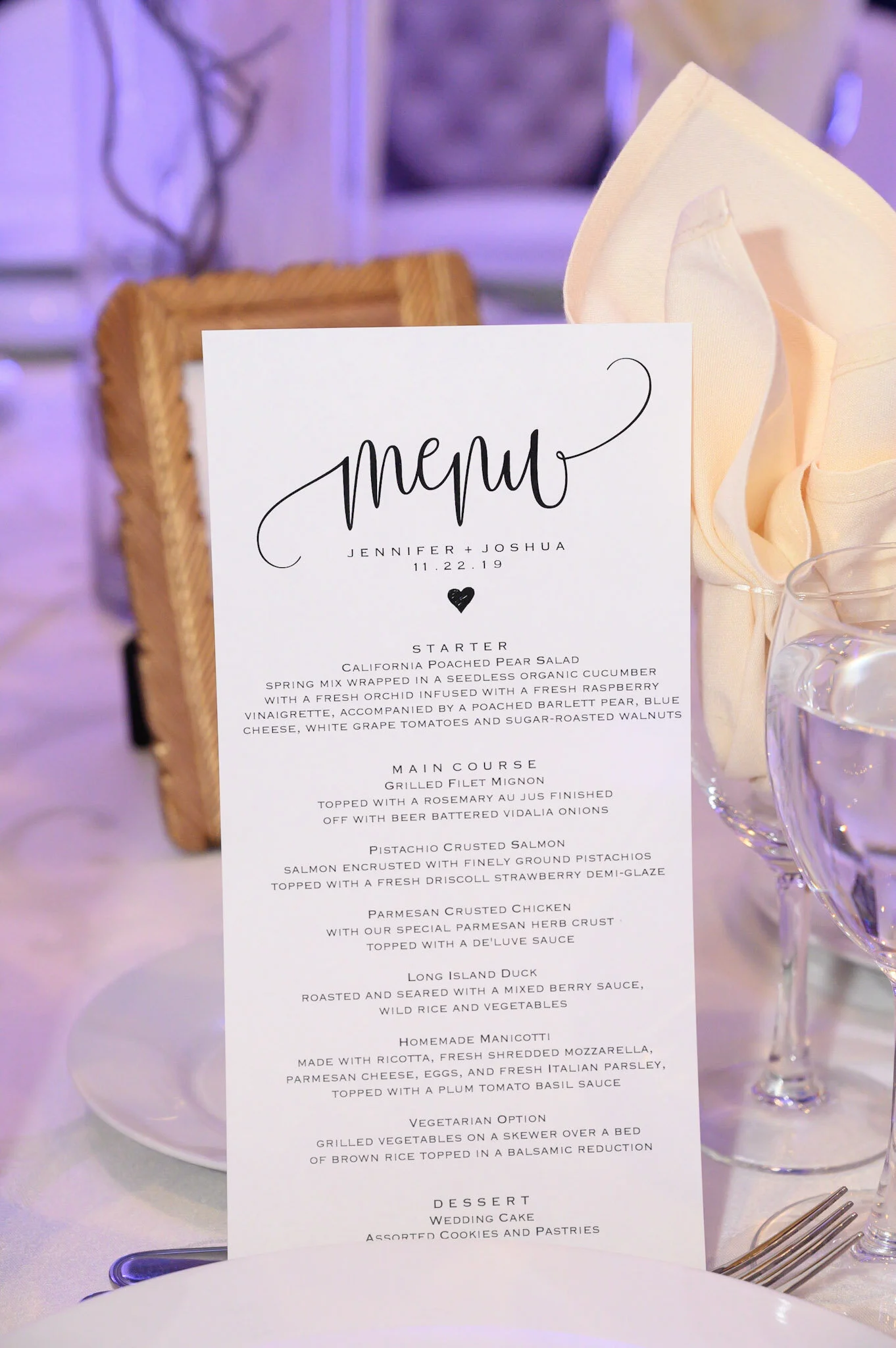 A wedding menu placed on a table with a background of blurred purple and white decorations. The menu is on white paper with elegant black handwritten and printed text, listing the courses for the wedding meal.