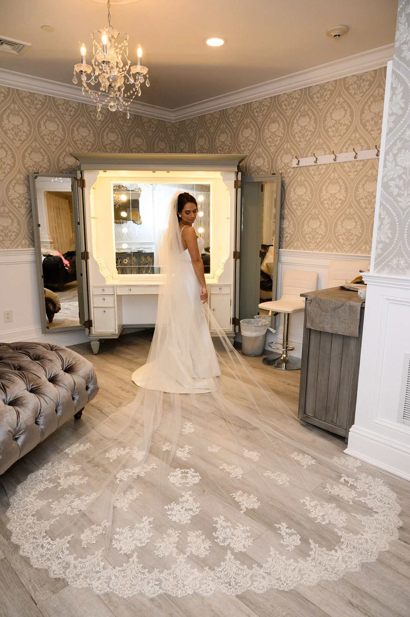 A bride in a wedding dress with a long lace veil standing in a well-lit bridal suite with a chandelier, chairs, and wallpapered walls.