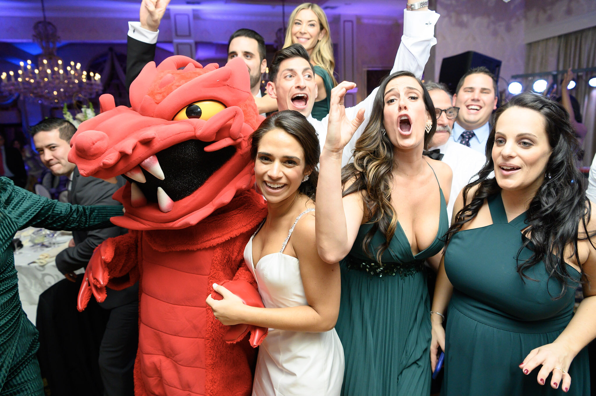 A group of people at a celebration, including a woman in a white dress, a woman in a teal dress, and a person in a red dragon costume with a large head and yellow eyes. The group appears excited and happy, with some raising their hands and shouting. 