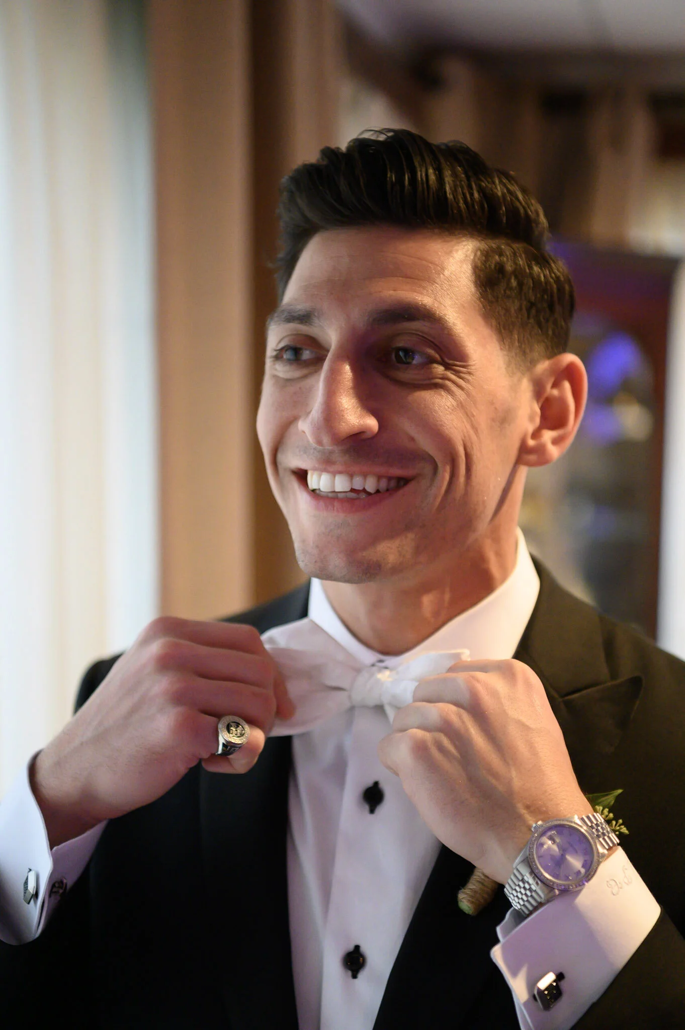 A young man in formal attire, adjusting a white bow tie, smiling, wearing a black tuxedo, wristwatch, and ring, with warm indoor lighting in the background.
