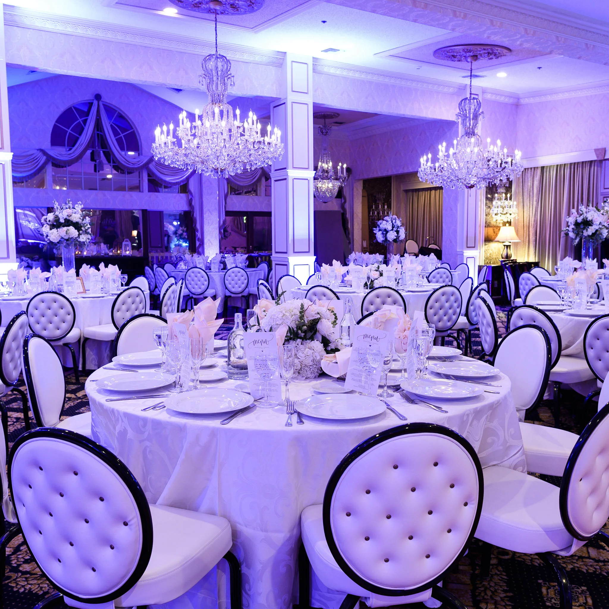 Elegant banquet hall decorated with white and lavender colors, illuminated by multiple large crystal chandeliers, with round tables set with white tablecloths, floral centerpieces, wine glasses, and place settings, and surrounded by white chairs with