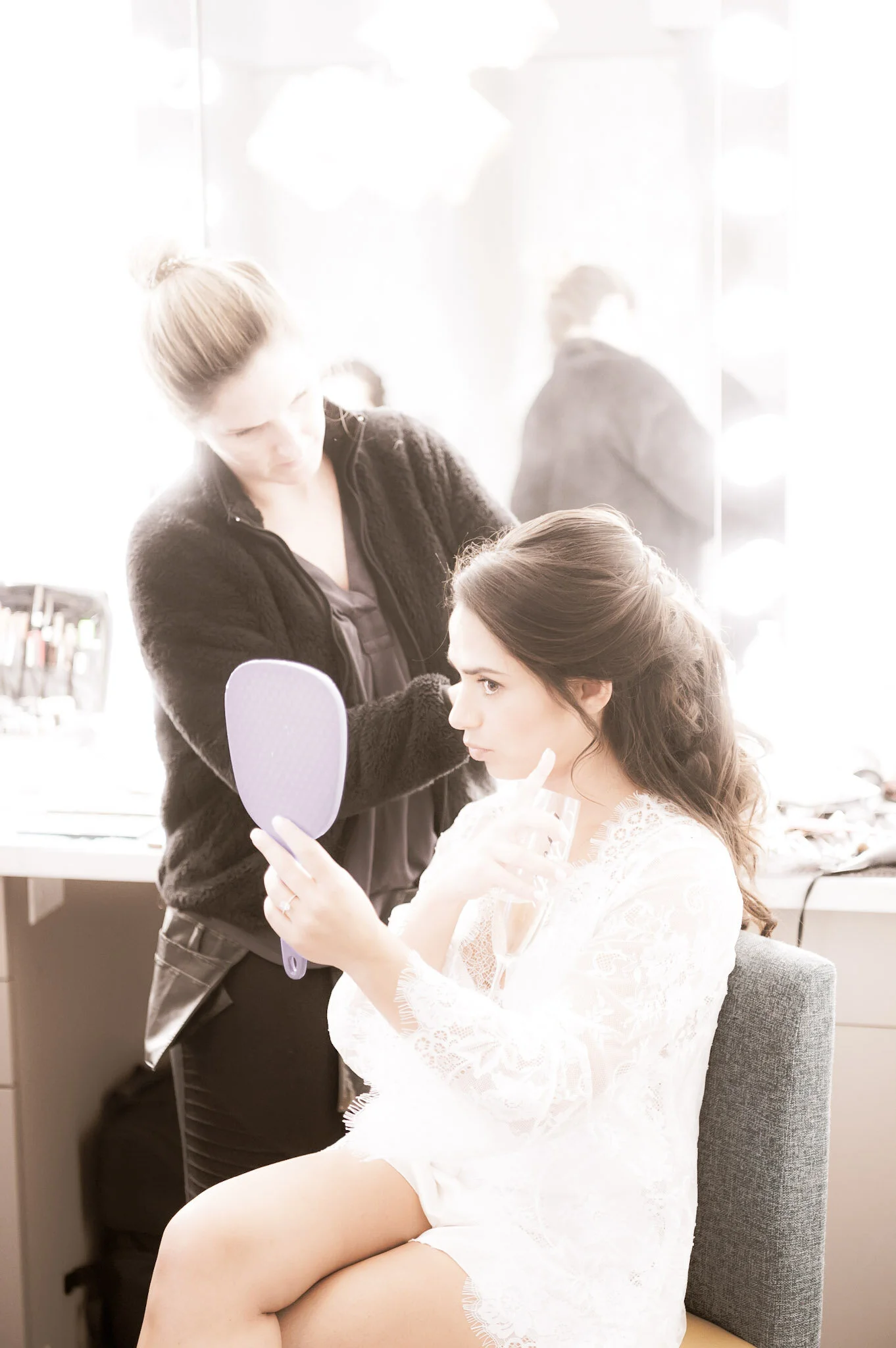 A woman with dark hair wearing a white lace robe sits in a salon chair, holding a mirror and looking at her reflection while a stylist with blonde hair in a bun, dressed in black, applies makeup or styling products to her face or hair. The background