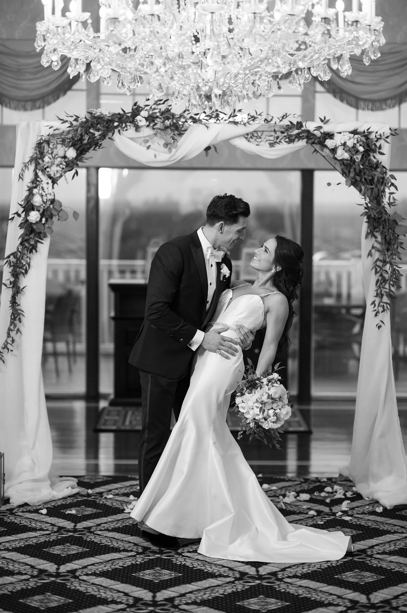 A bride and groom are exchanging vows under a floral arch and crystal chandelier during a wedding ceremony, with a patterned rug and a bouquet of flowers in the bride's hand.