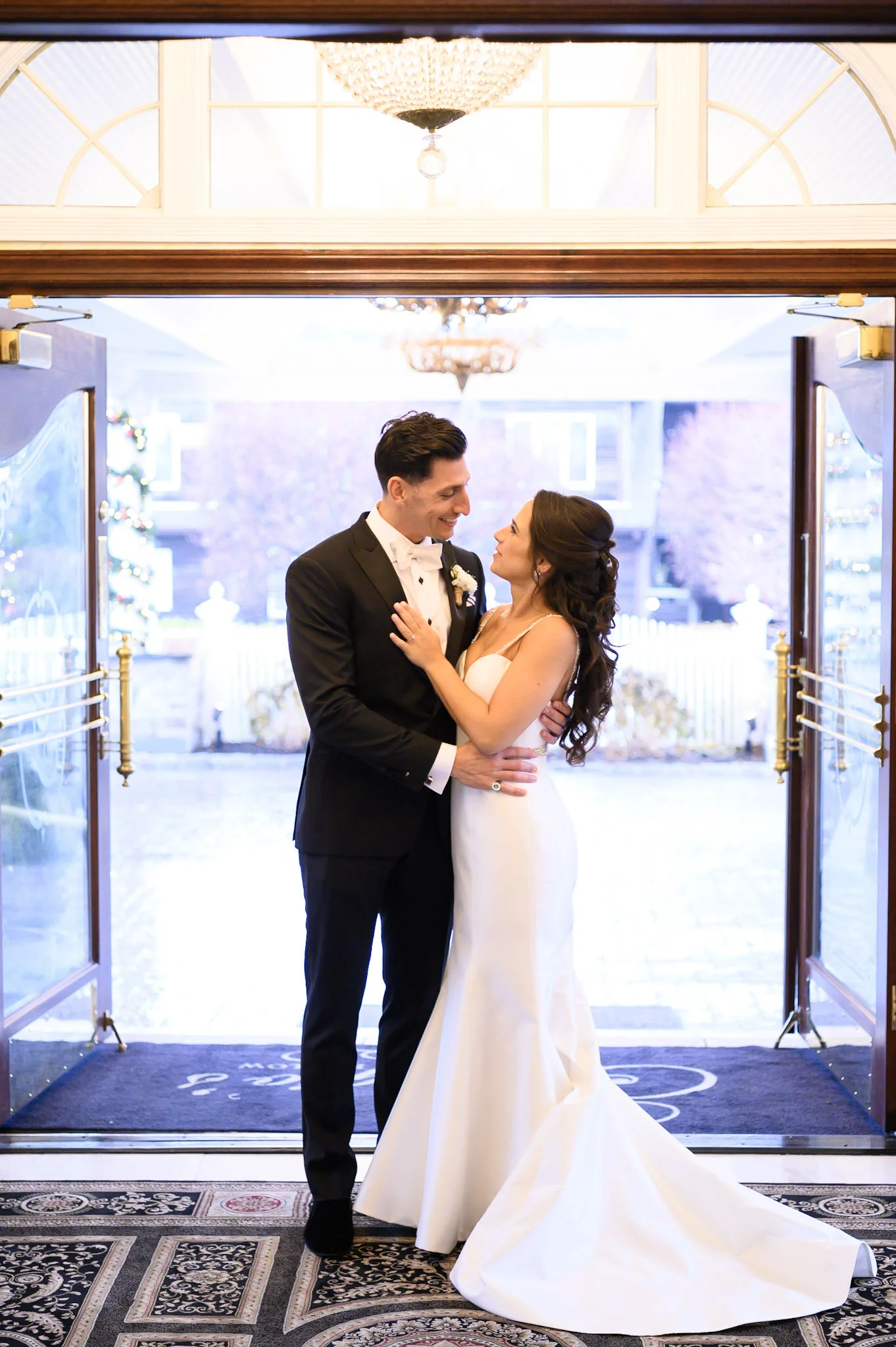 A bride and groom share a moment at the entrance of a wedding venue. The groom wears a black tuxedo with a white shirt and bowtie, while the bride wears a white gown with long dark hair. The background features a decorated outdoor area with Christmas