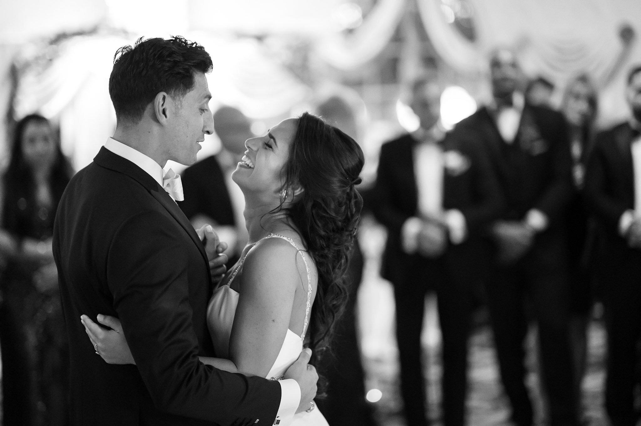 A couple dancing at a formal event, with the man wearing a suit and tie and the woman in a gown, smiling at each other, with other guests in the background.