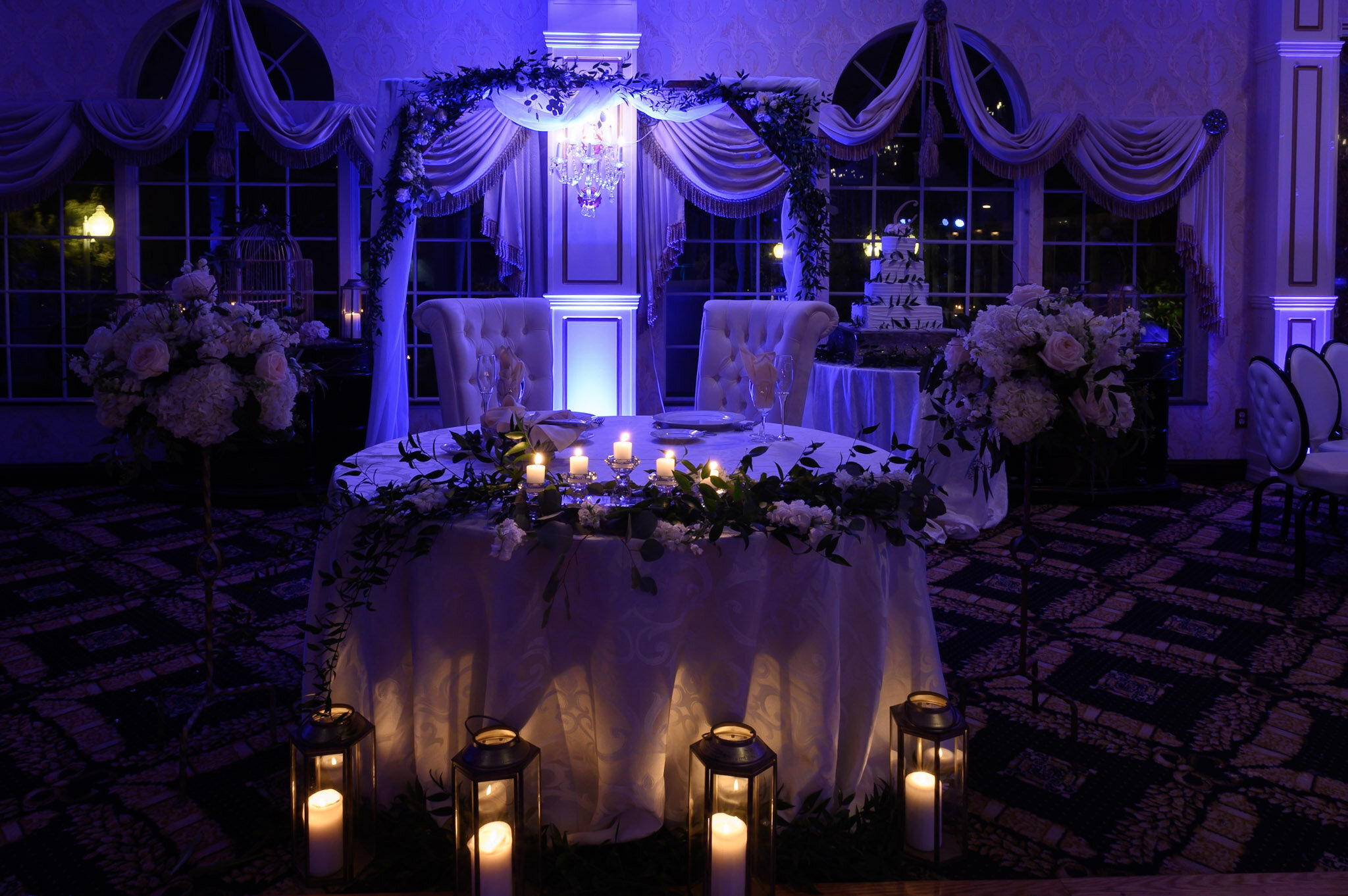 Elegant wedding reception decorated with floral arrangements, candles, draped fabric, and a tiered wedding cake in a dimly lit room with blue lighting.