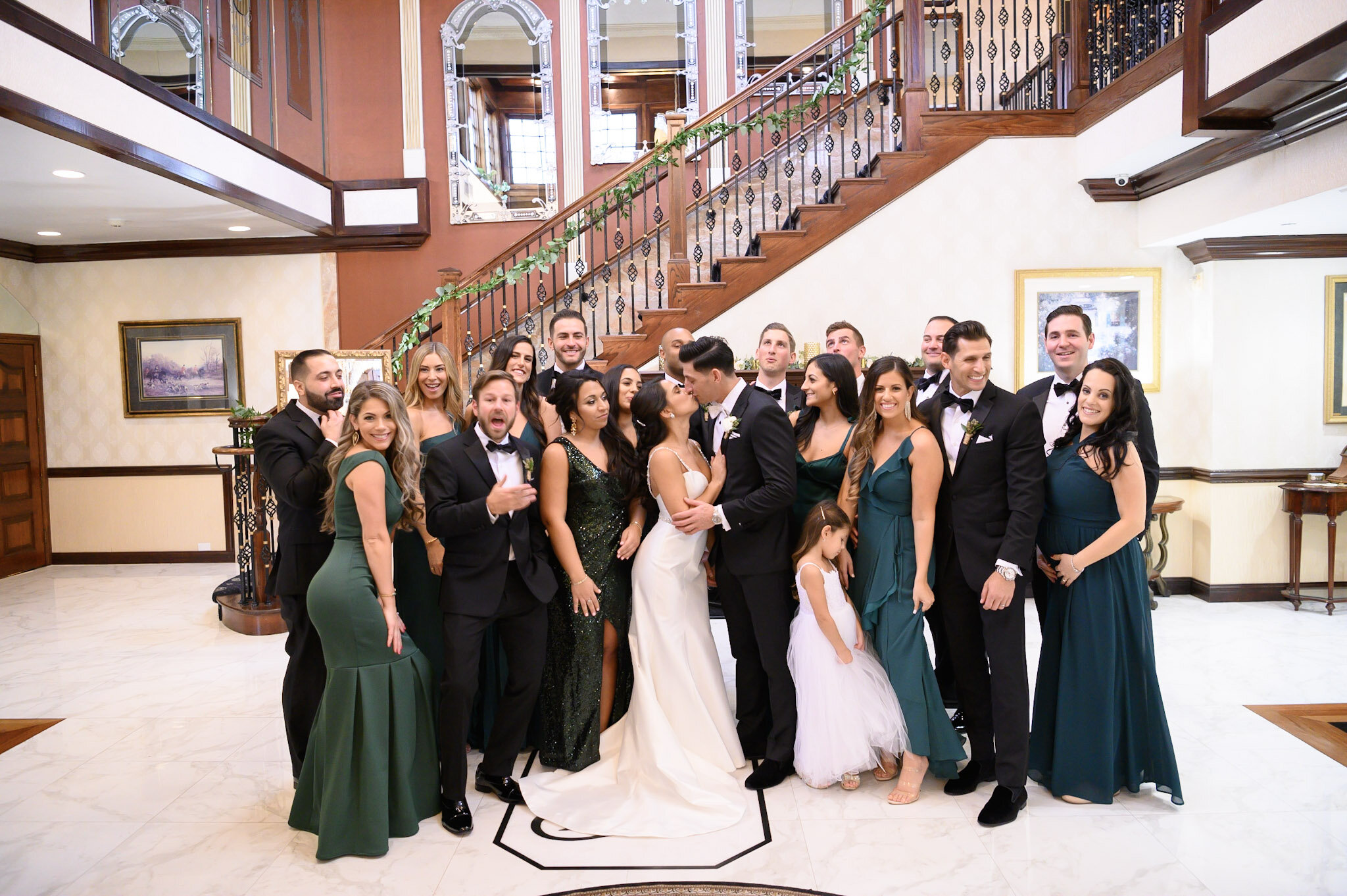 Group of people at a wedding reception, including bride and groom, dressed in formal attire, standing indoors in front of a staircase with wooden railing and decorative mirrors.