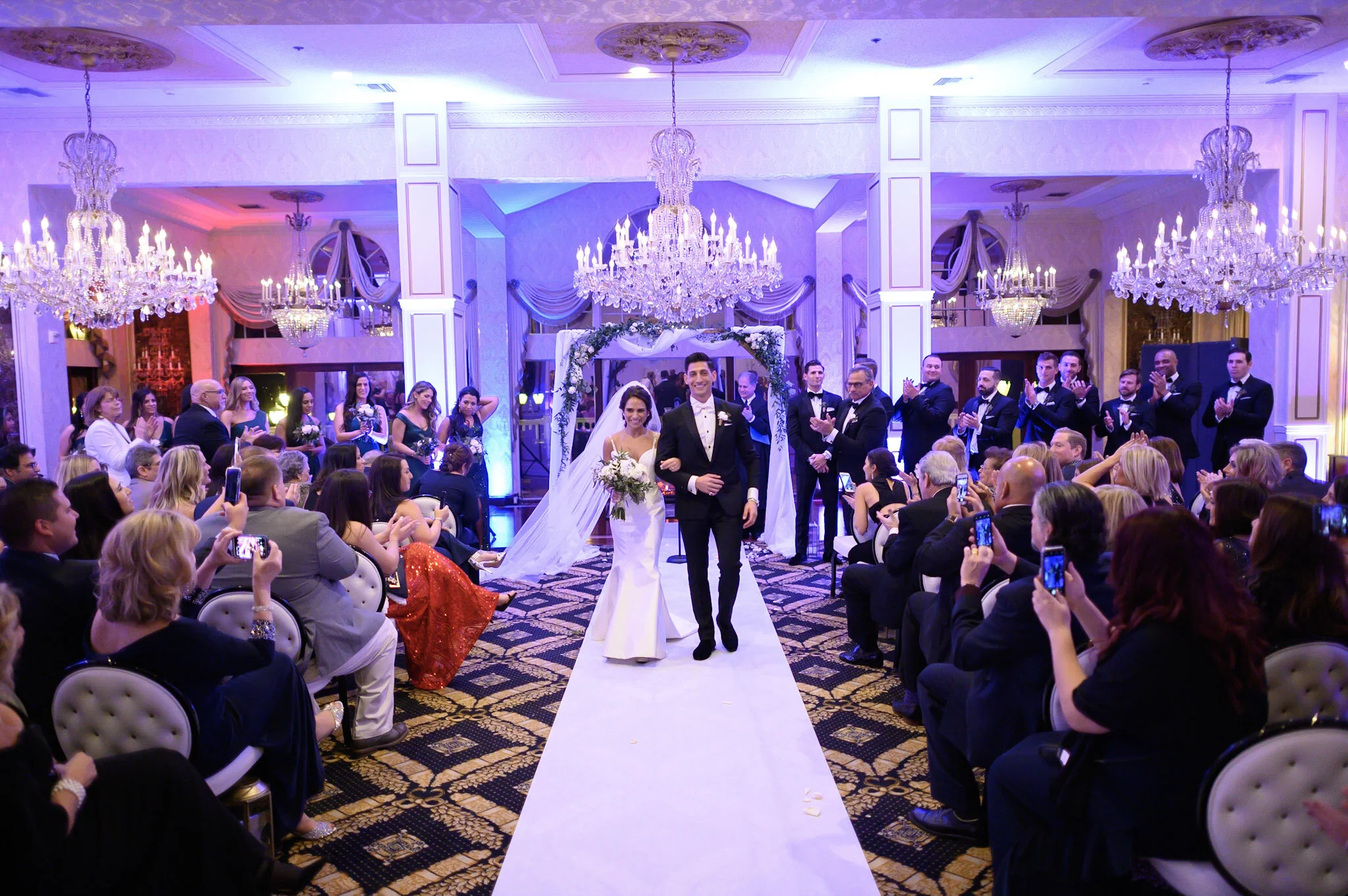 A wedding ceremony with the bride and groom walking down the aisle in a banquet hall decorated with chandeliers and drapery, surrounded by seated guests and wedding party.