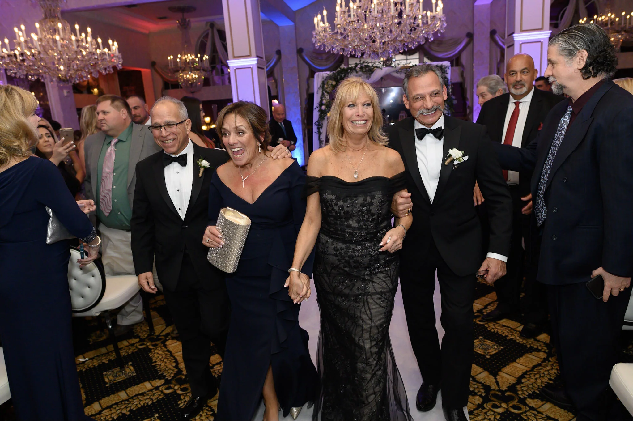A group of people dressed in formal evening attire, socializing and having fun at an elegant event with chandeliers and purple lighting.