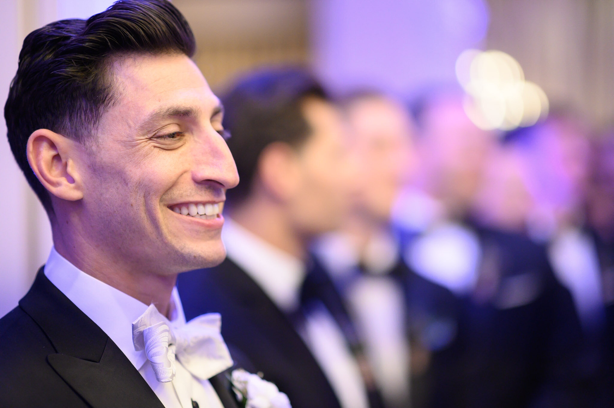 A man in a tuxedo with a white bow tie smiling at an event, with a blurred background of other people also dressed formally.