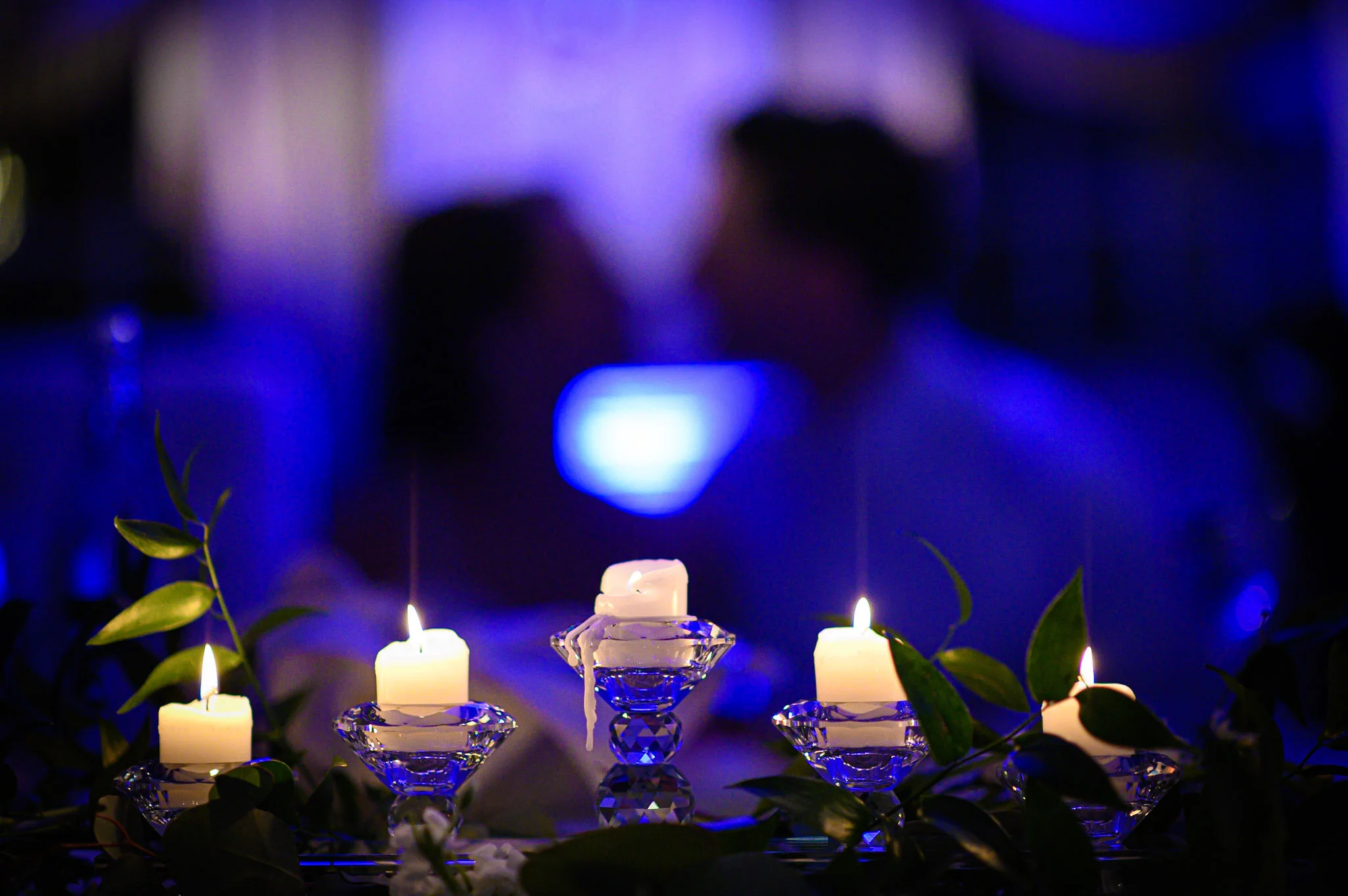 Multiple lit candles arranged in glass holders with green leaves around them, set against a dark blue and purple background.