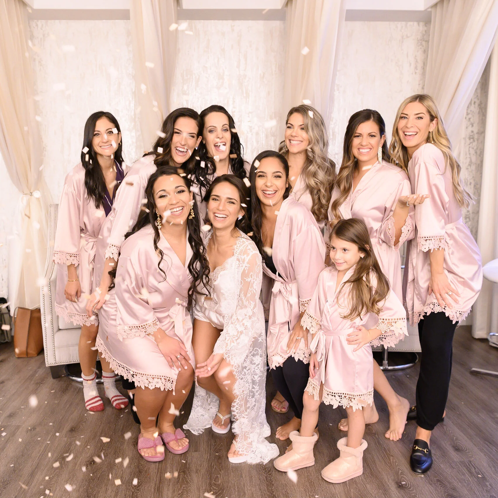 A group of women, dressed in matching pink robes, gathered together and celebrating with confetti. They are smiling and appear to be at a bridal shower or similar celebration.