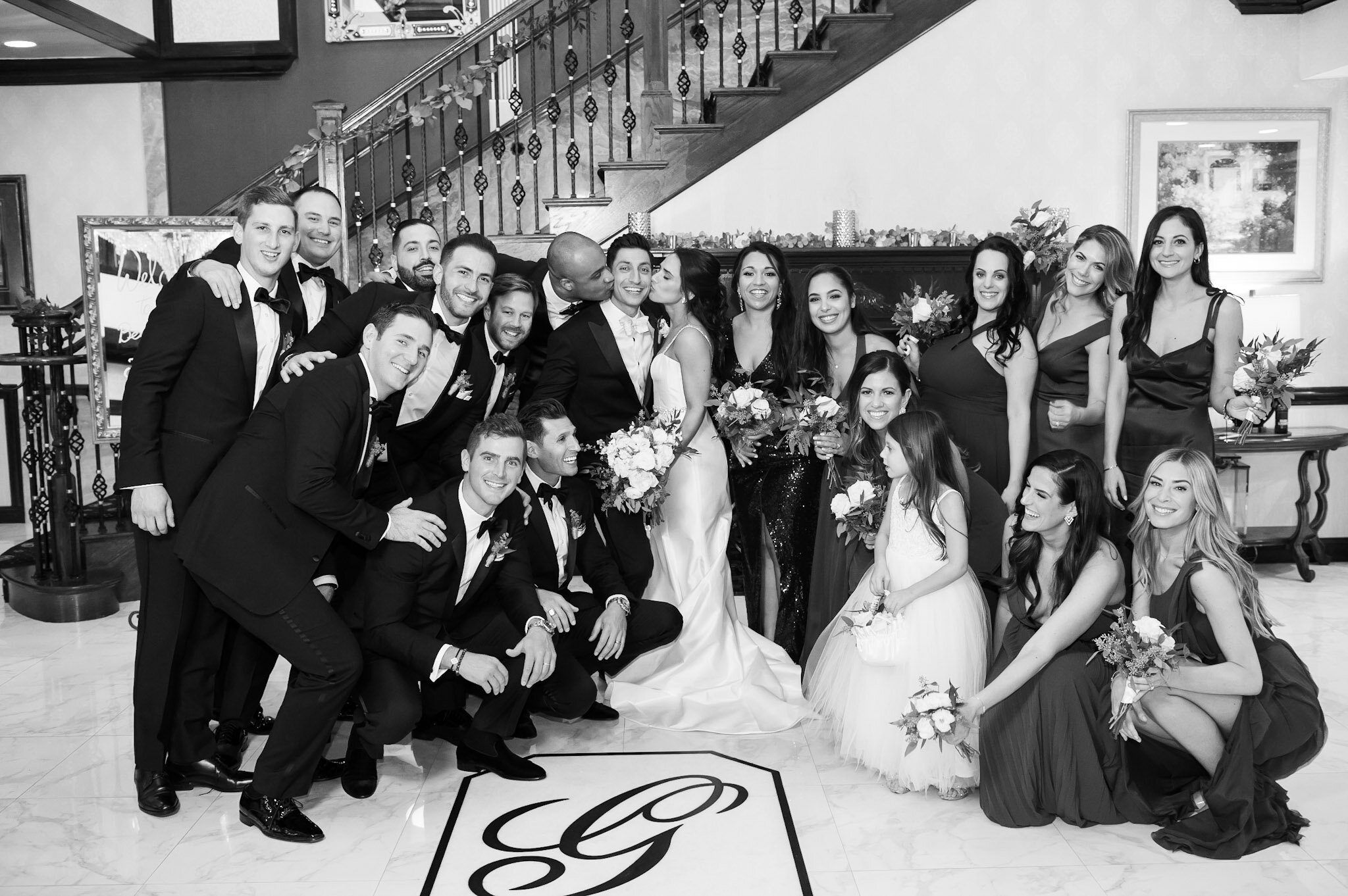 A group of men and women in formal attire, probably at a wedding, standing together indoors in front of a staircase, smiling and posing for the photo. The women are holding bouquets, and the men are wearing tuxedos.