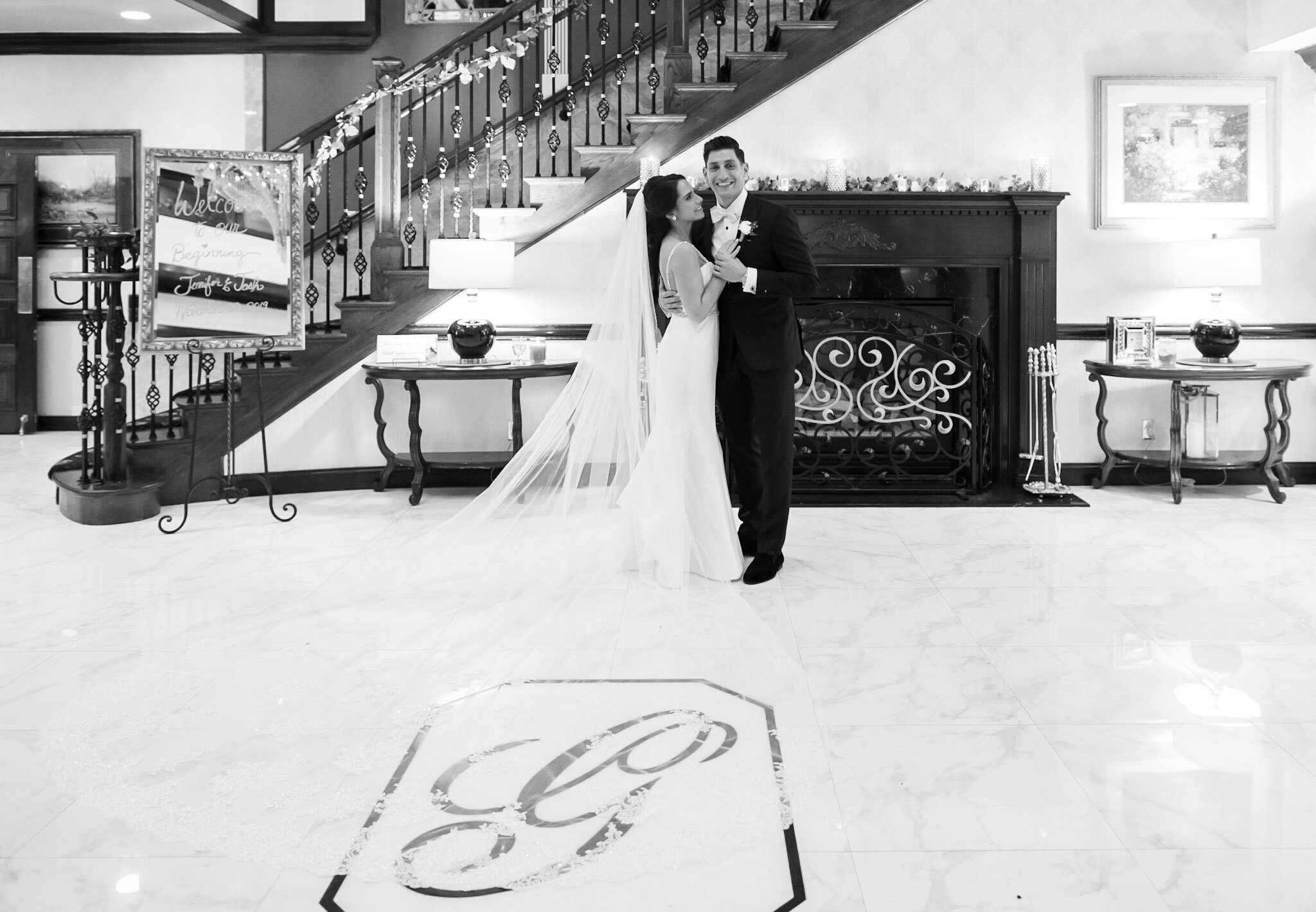 A bride and groom dancing indoors, with the bride in a white wedding gown and veil, and the groom in a black tuxedo. They are smiling and holding each other close, under a staircase decorated with flowers and candles, in a warmly lit room with artwor