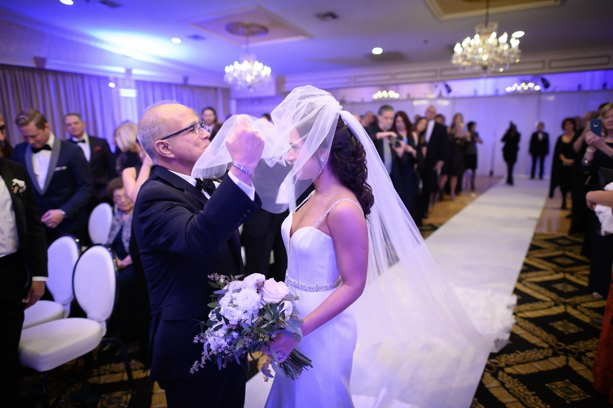 A bride and an older man, possibly her father, sharing a moment during a wedding reception in a decorated ballroom with chandeliers. The older man is adjusting the bride's veil.