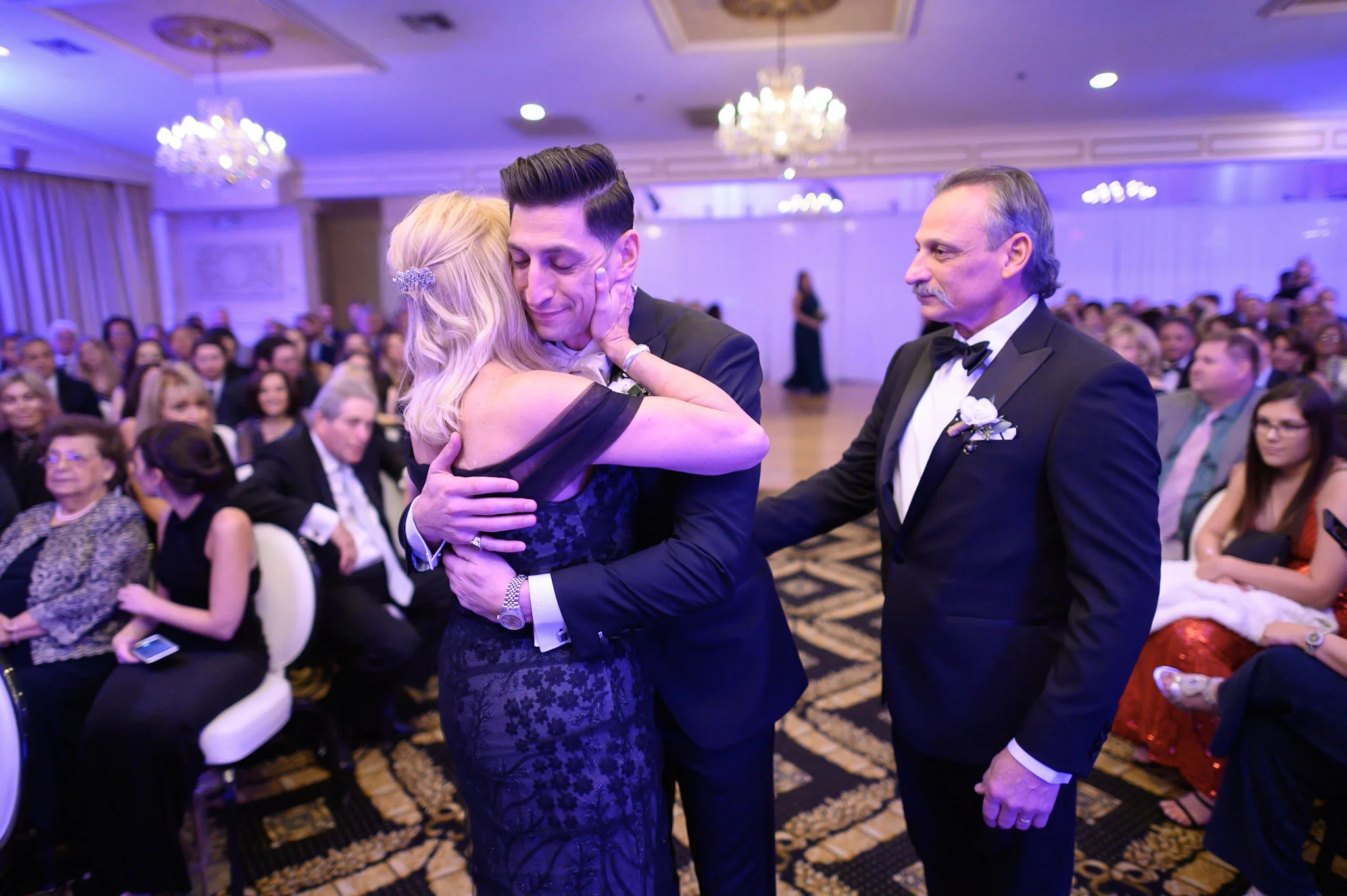 A man in a tuxedo and a woman in a black lace dress embrace during a dance at a formal event, with people seated in the background.
