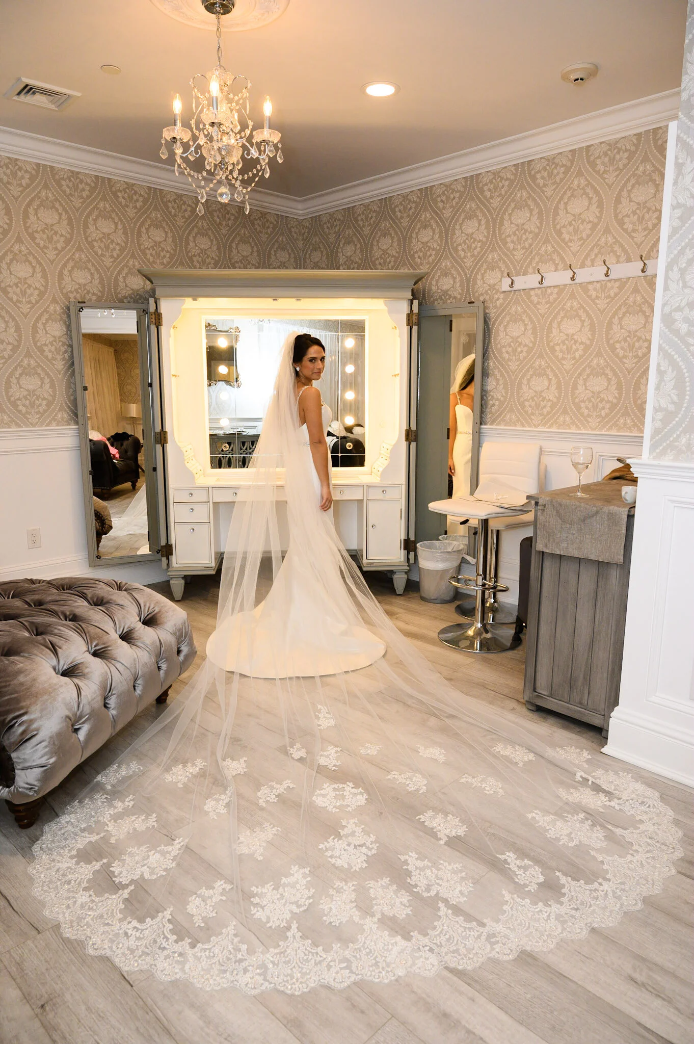 A bride in a white wedding gown with a long veil standing in front of a vanity mirror with lights. The room has elegant wallpaper, a chandelier, a tufted ottoman, a white chair, and a lace-trimmed wedding train on the floor.