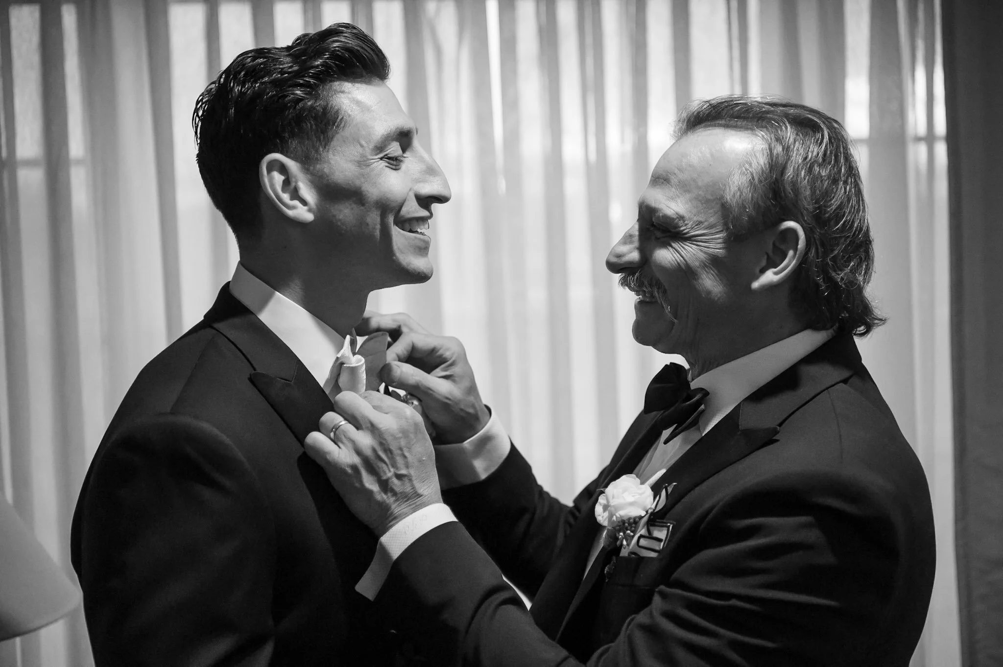 Two men, one younger and one older, dressed in tuxedos, sharing a joyful moment, smiling, in front of a curtain.