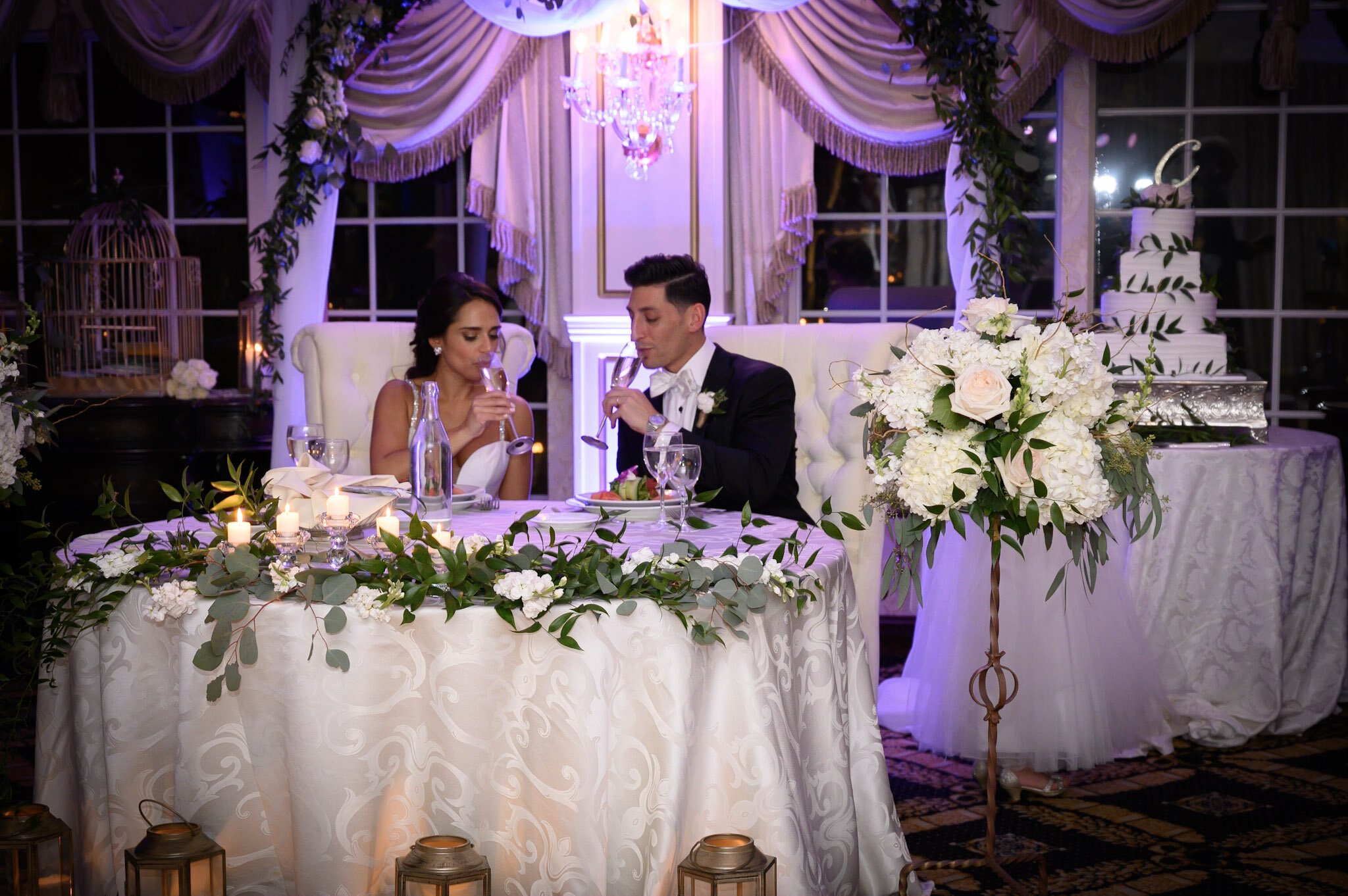 A bride and groom sitting at a decorated wedding reception table with floral centerpieces, candles, and elegant decorations in a lit ballroom.