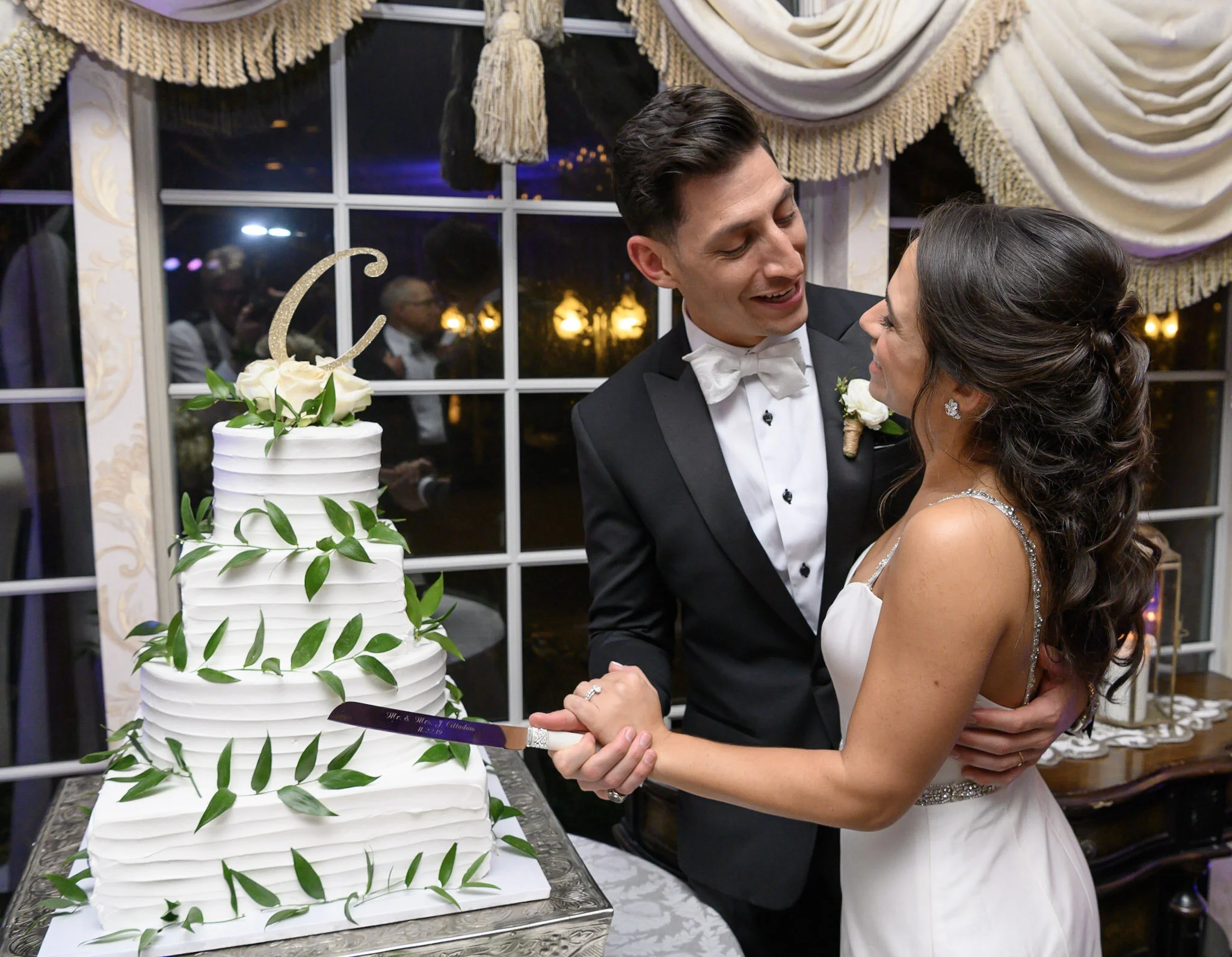 A newlywed couple, a groom in a tuxedo and a bride in a wedding gown, share a moment of joy and affection during their wedding celebration. They are holding hands and looking at each other near a multi-tiered white wedding cake decorated with greener