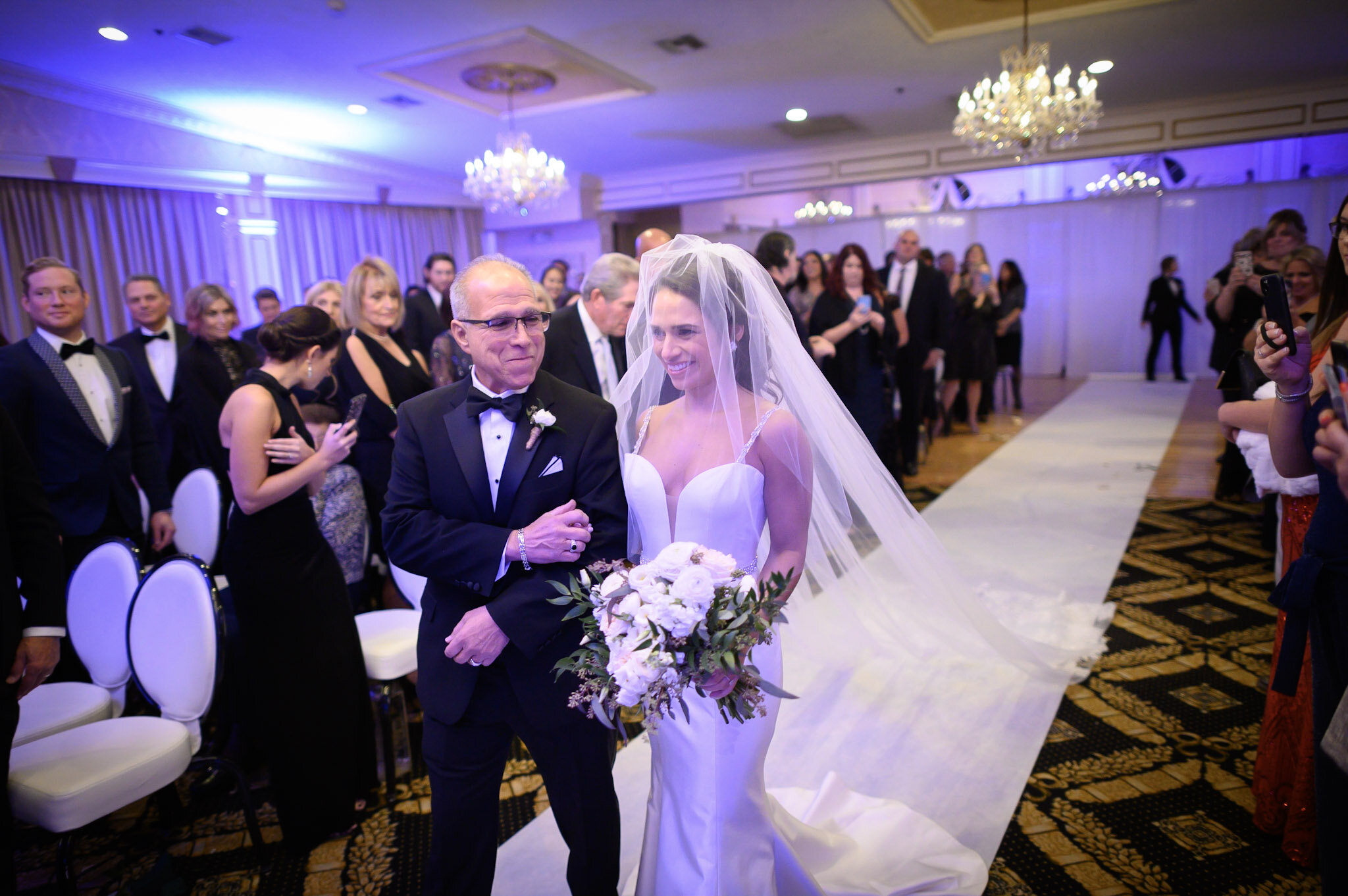A bride and an older man, possibly her father, are walking down the aisle at a wedding ceremony. The bride is wearing a white wedding gown and veil, holding a bouquet of flowers. The man is dressed in a black tuxedo with a bow tie. Guests are seated 