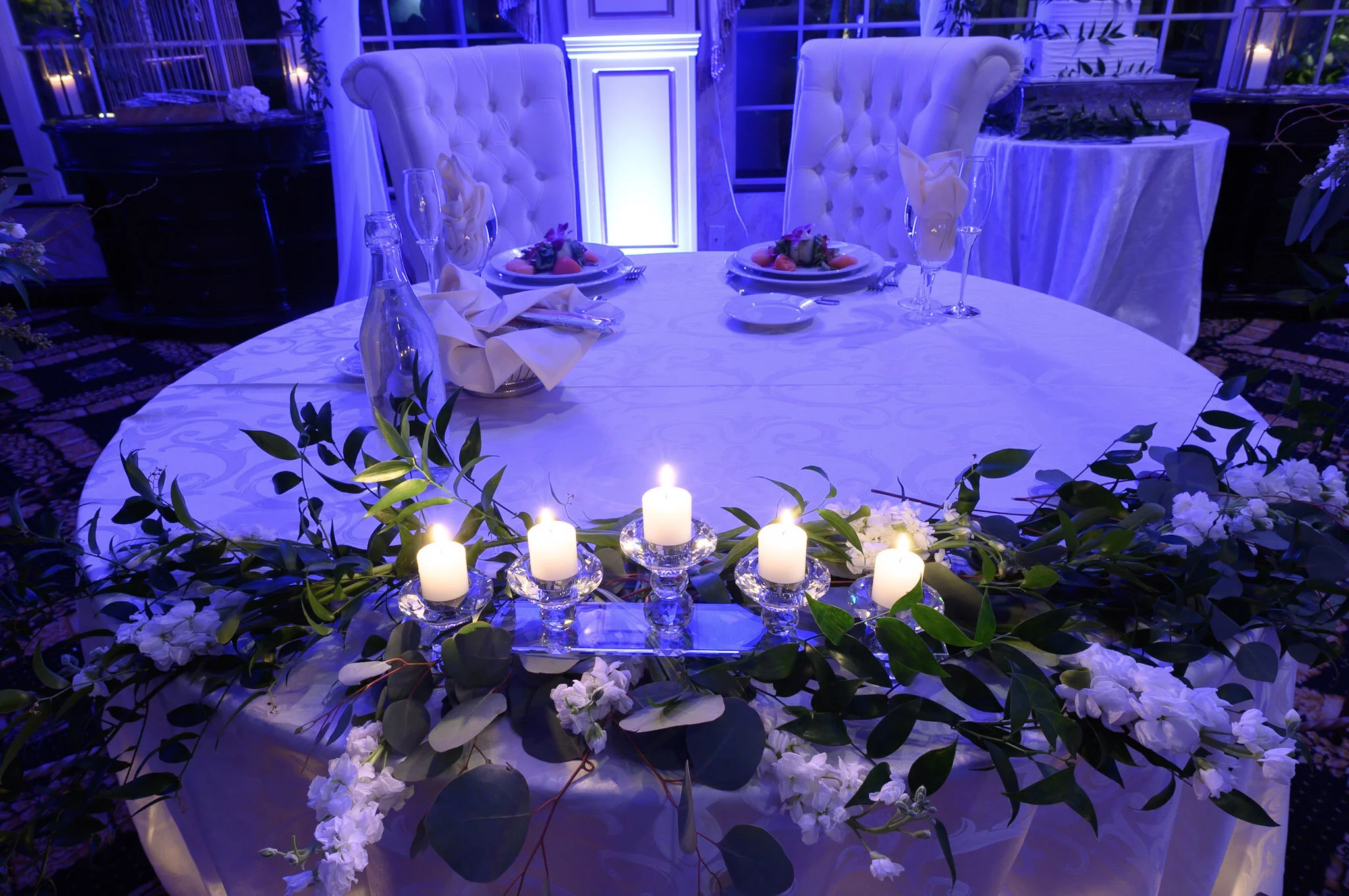 A table set for a special occasion, decorated with white flowers, green leaves, and lit candles. The background features two white tufted chairs, and blue lighting enhances the elegant ambiance.