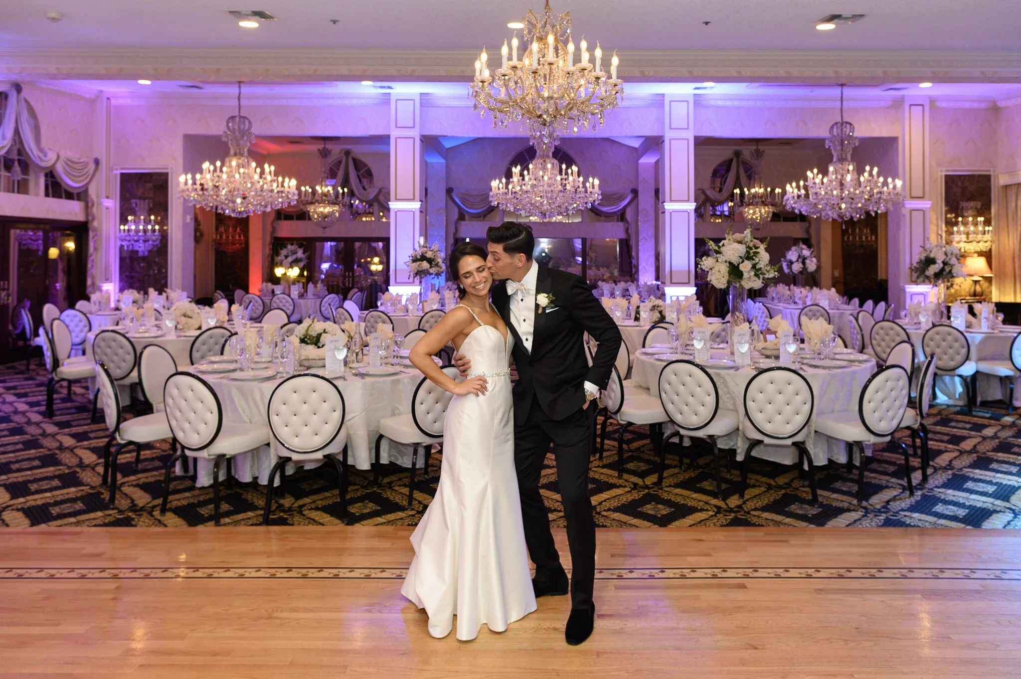 A bride and groom dancing in a decorated wedding reception hall with chandeliers, round tables, and floral arrangements.