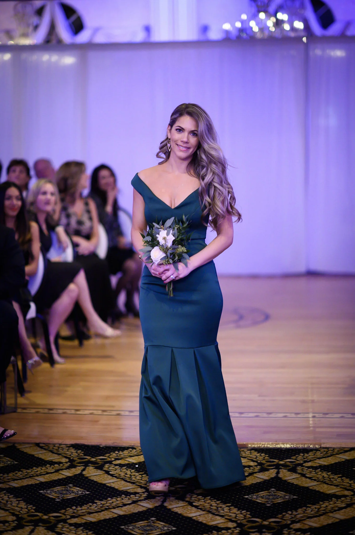 A woman in a dark teal dress holding a small bouquet of flowers at a formal event with seated guests in the background.